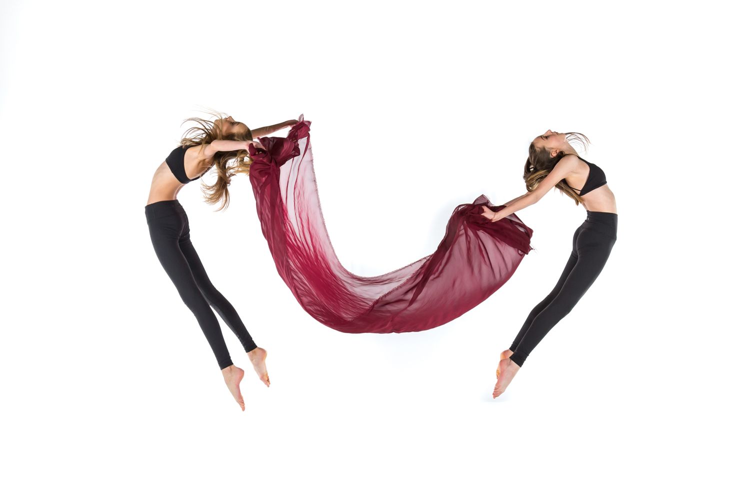 Two dancers in black leggings gracefully leap while holding a flowing burgundy fabric between them against white background.
