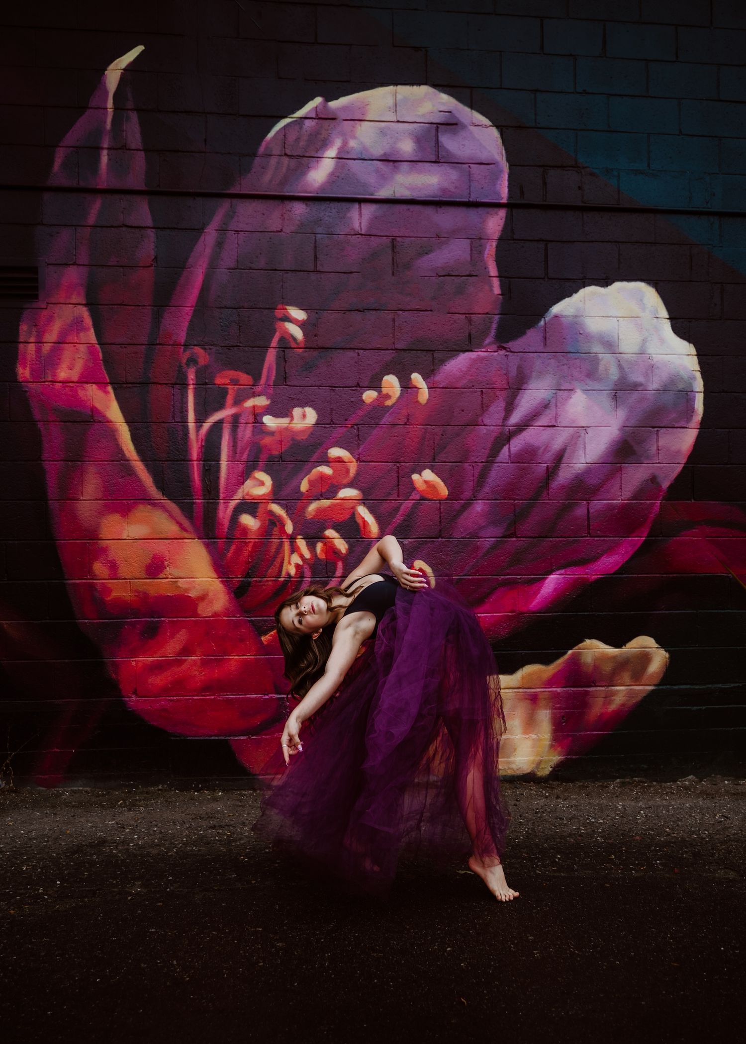 Dancer in purple dress poses against vibrant floral street art mural with dramatic lighting.