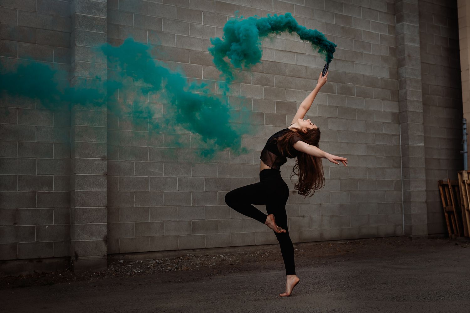 A dancer in black leaps gracefully against a brick wall while swirling a teal smoke bomb creating ethereal effects.