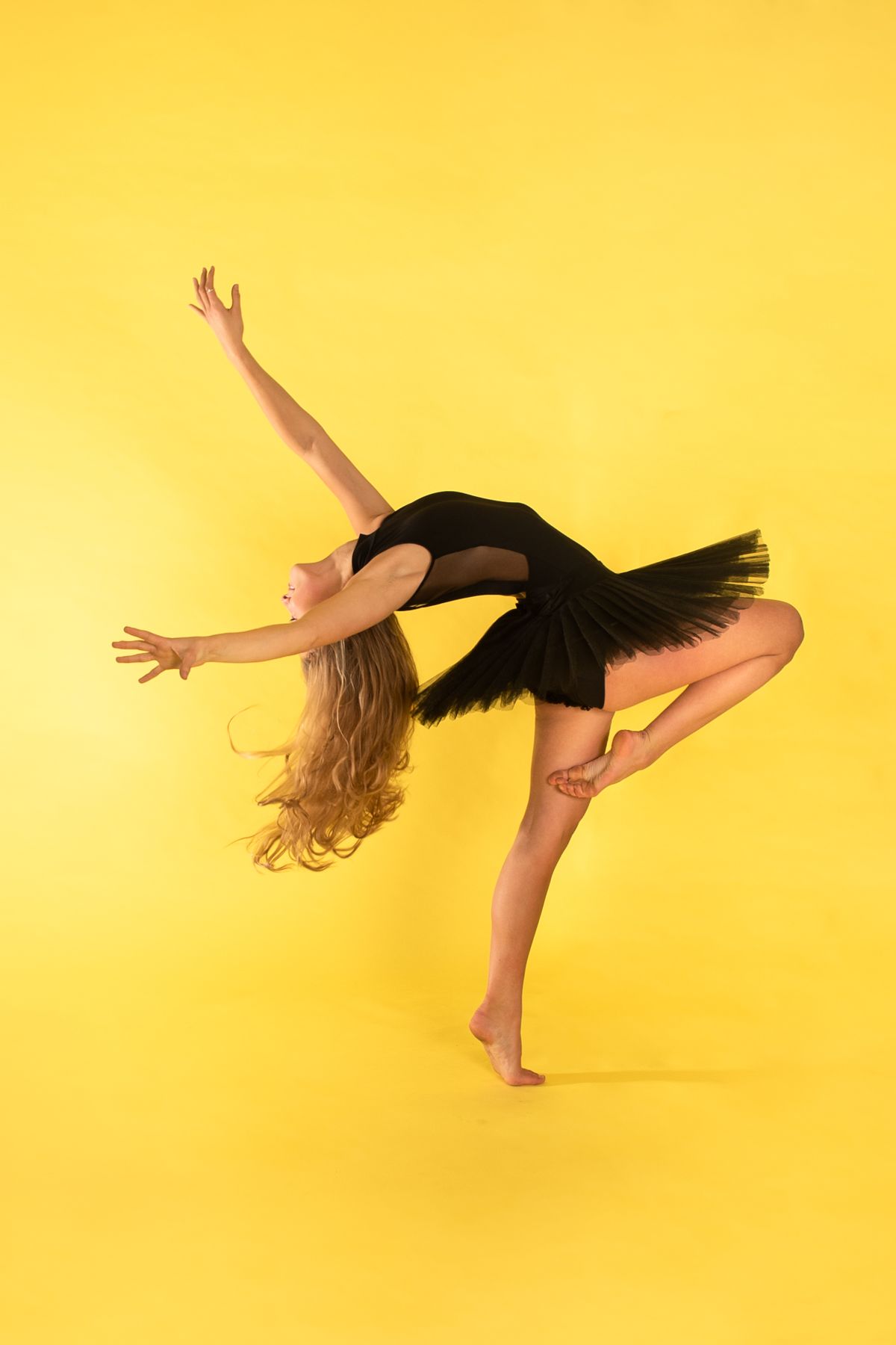 Modern dancer in black dress performs graceful pose against vibrant yellow backdrop, demonstrating elegant balance.