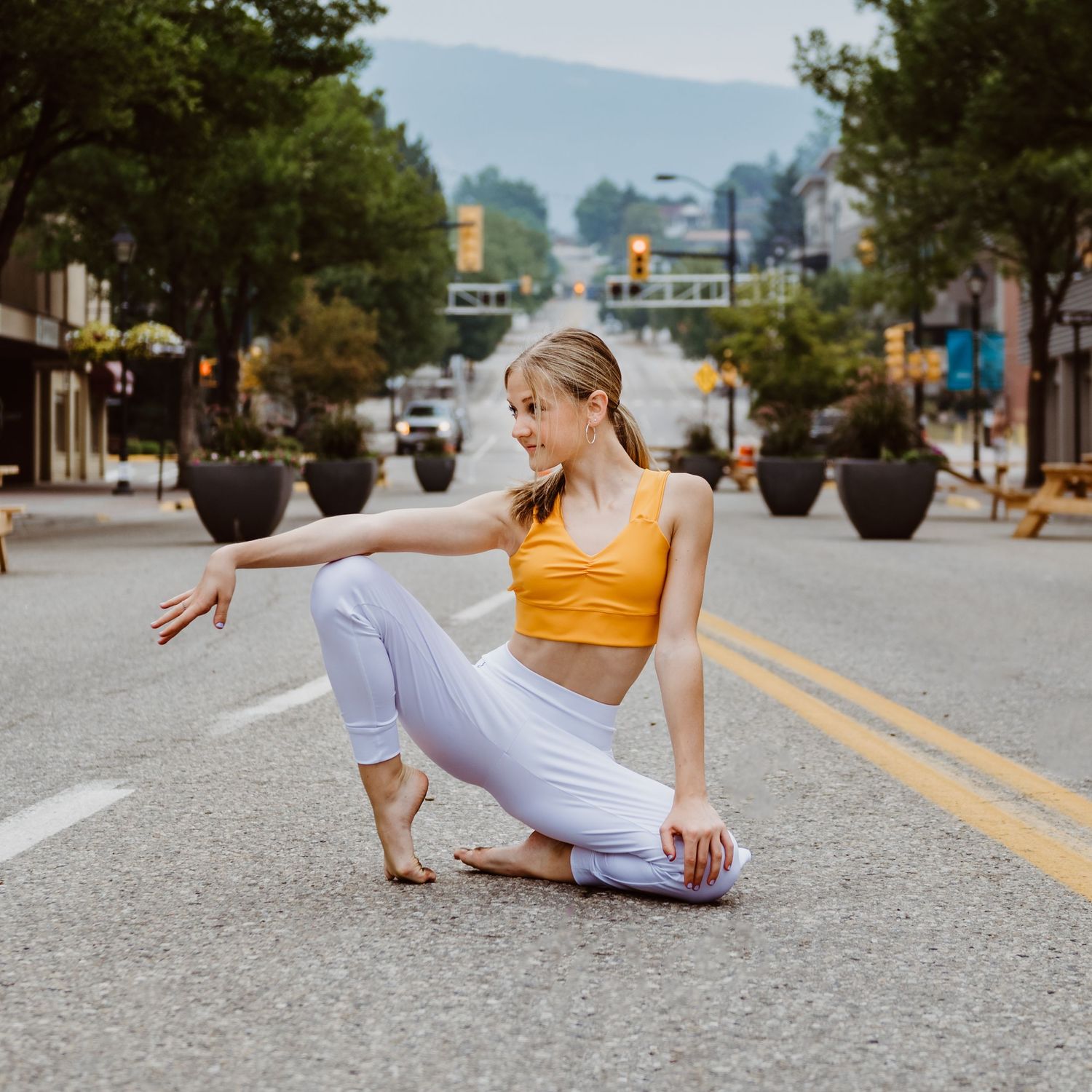 A dancer in bright yellow sportswear and white pants strikes a graceful pose in the middle of an urban street.