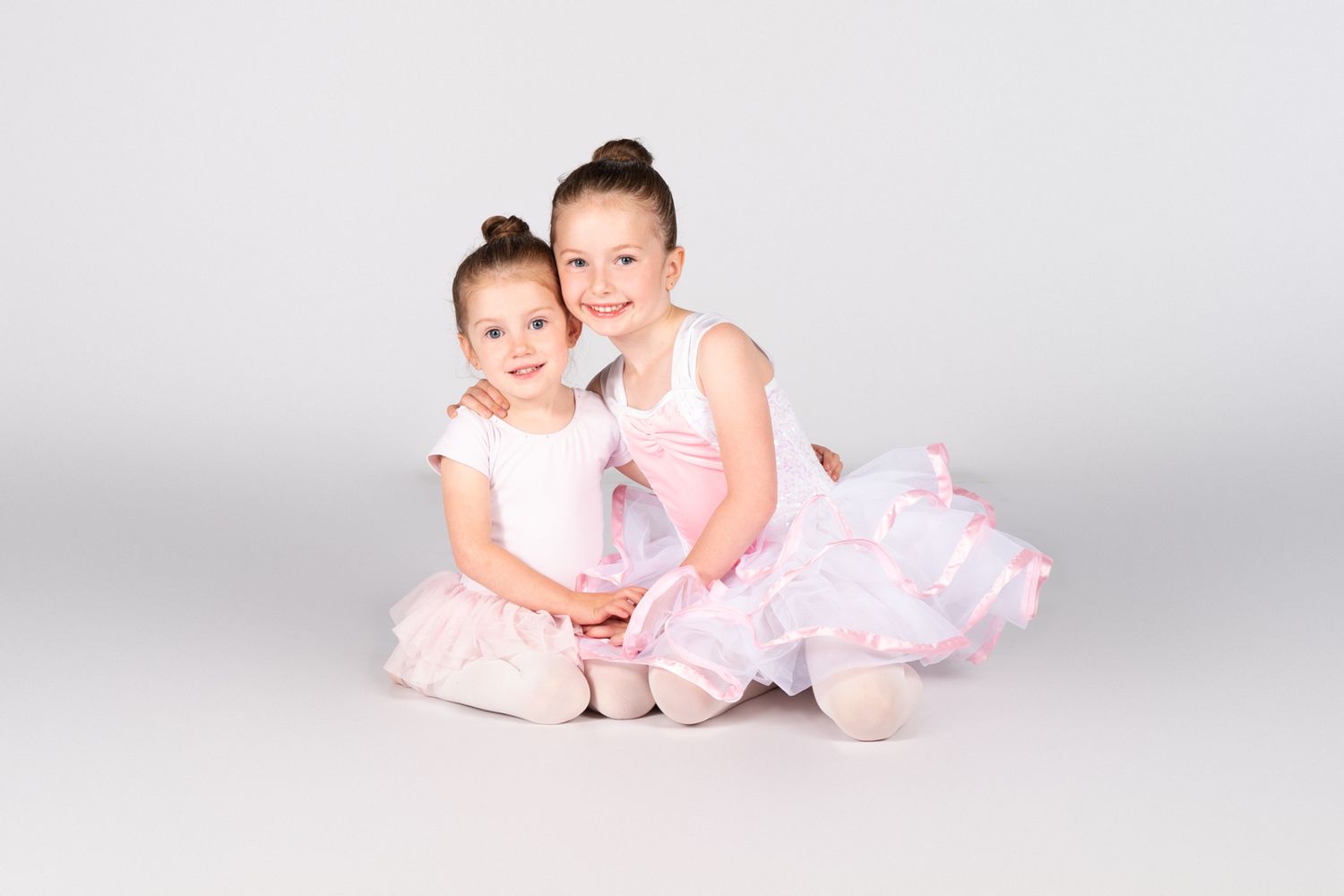 Two dancers wearing pink tutus pose together in studio against white background.