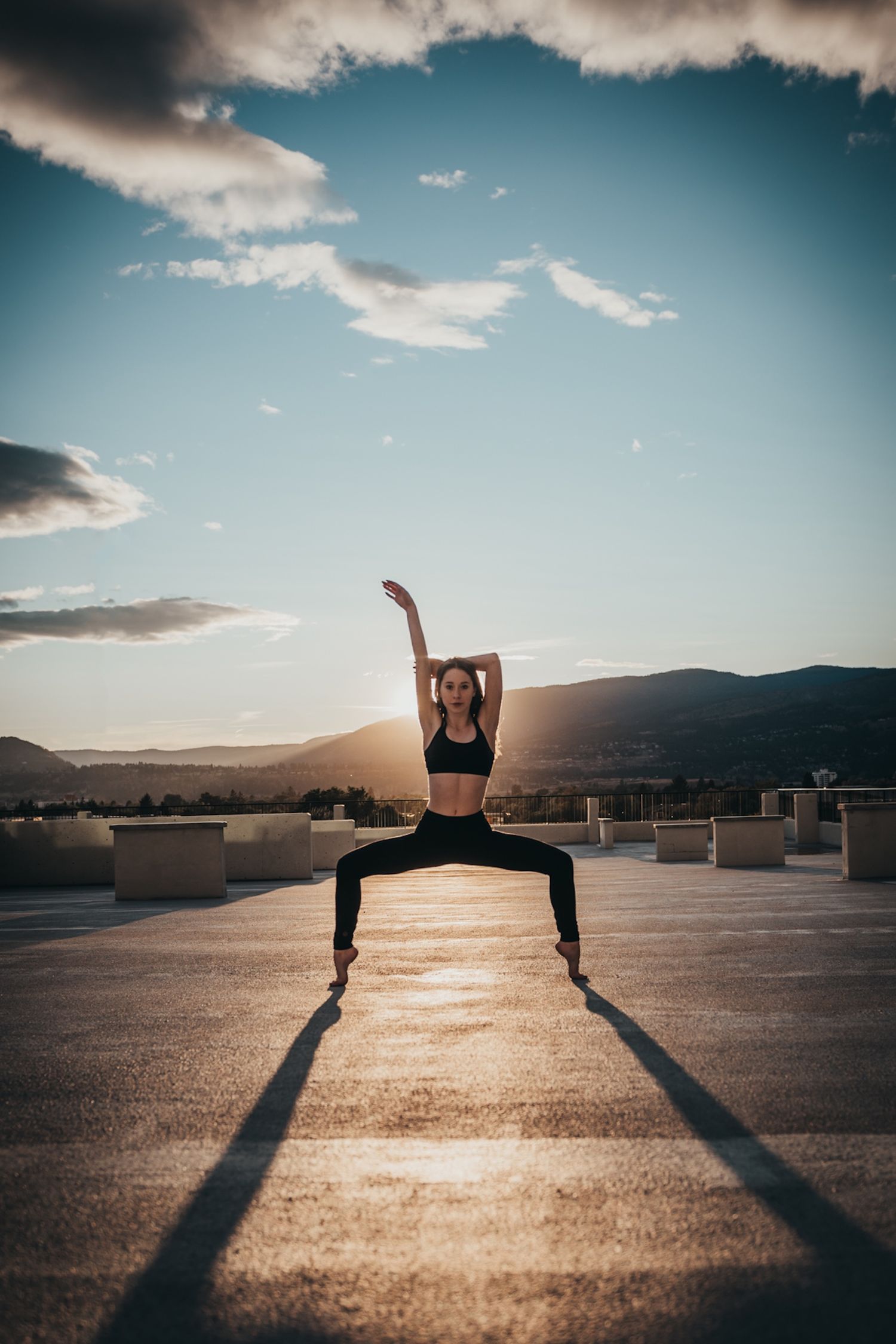 Silhouetted dancer performs athletic pose on rooftop at sunset with dramatic mountain landscape and cloudy sky backdrop.
