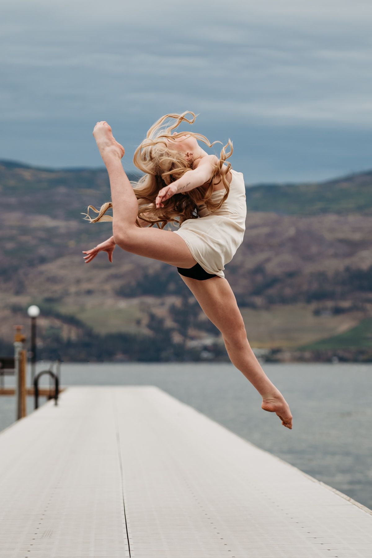 A dancer performs a graceful leap on a dock overlooking a lake and mountains on an overcast day.