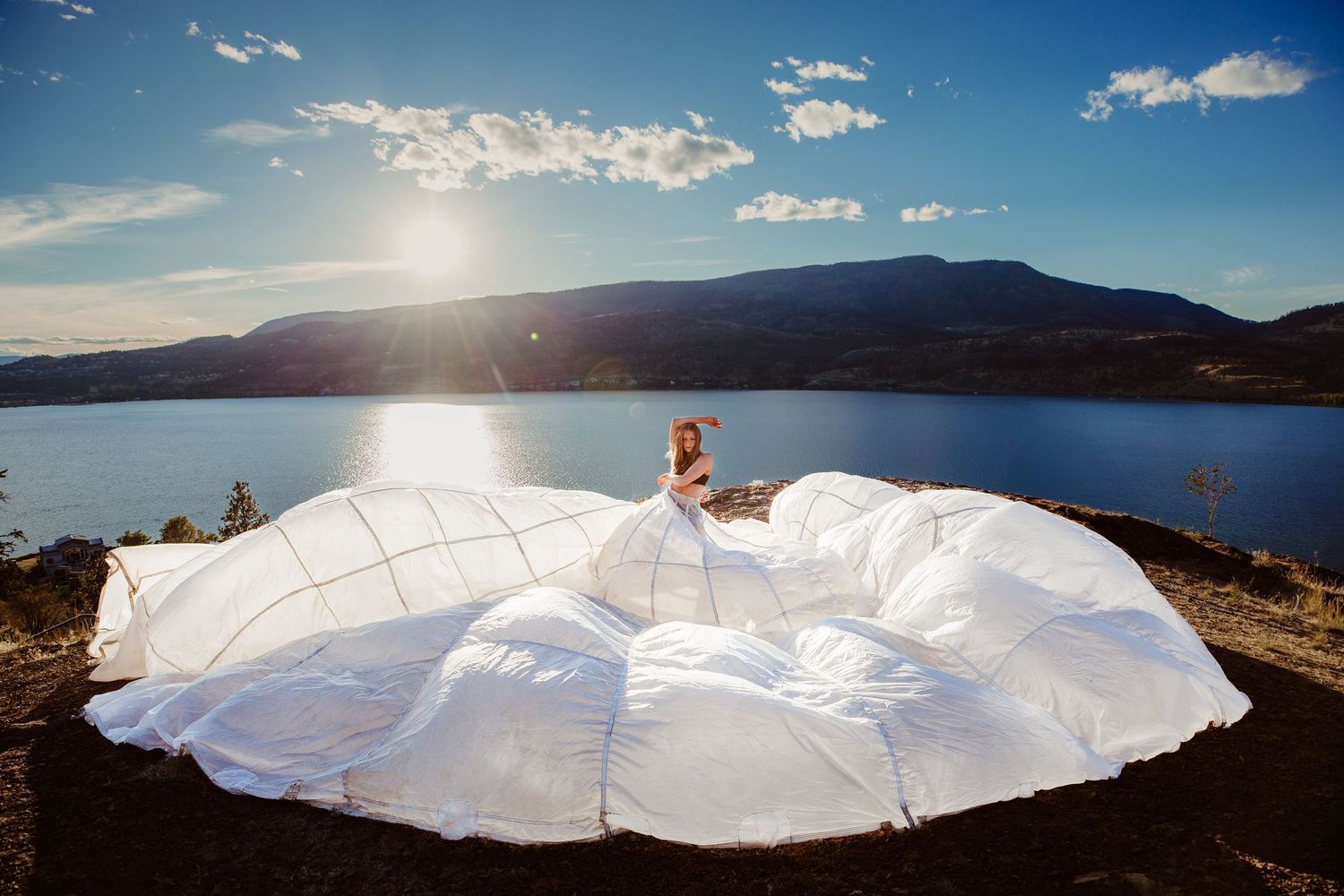 A flowing white wedding dress billows dramatically on a mountain overlook with a sunlit lake and blue sky in the background.