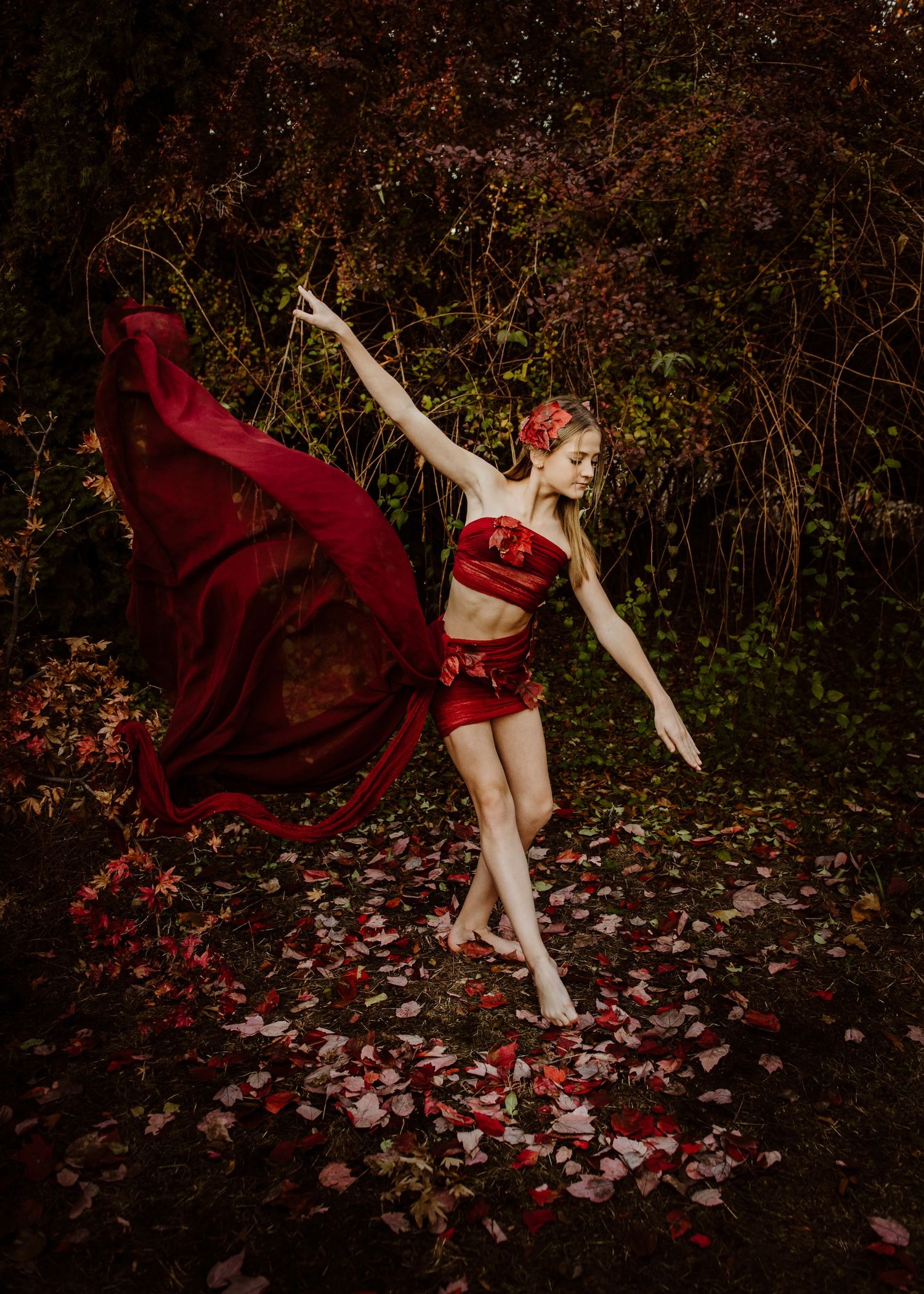 Dancer in flowing red costume performs graceful pose in autumn forest surrounded by fallen leaves.