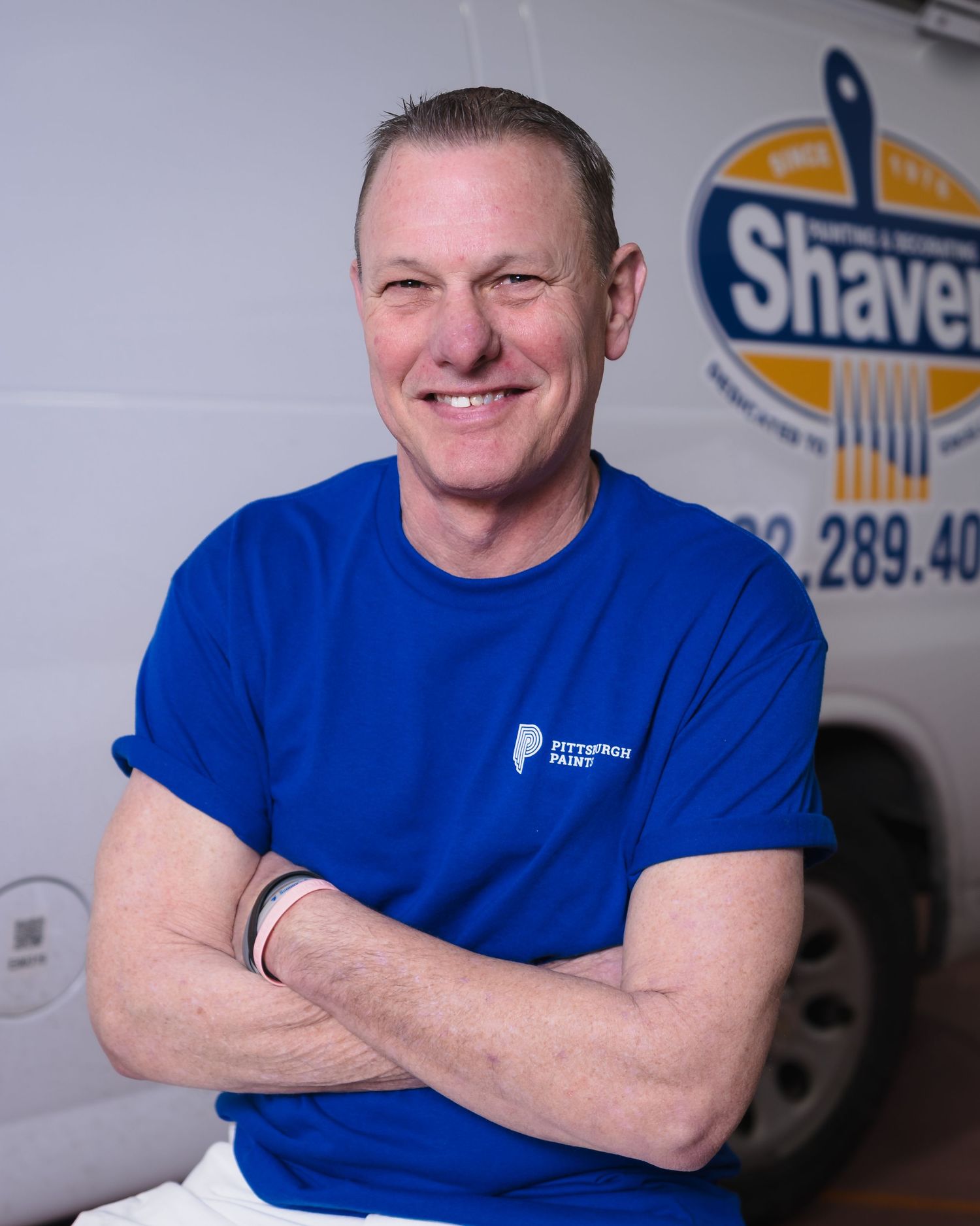 A smiling man in a blue Pittsburgh Paint shirt stands with arms crossed in front of a Shaver painting company van.