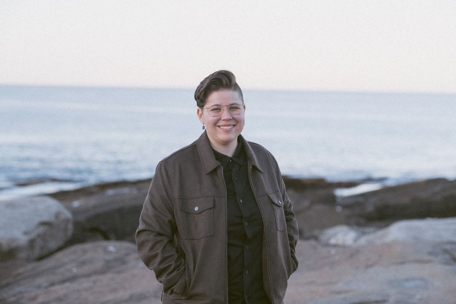 Person smiling outdoors on rocky coastal shoreline, wearing a brown jacket with ocean and sky in background.