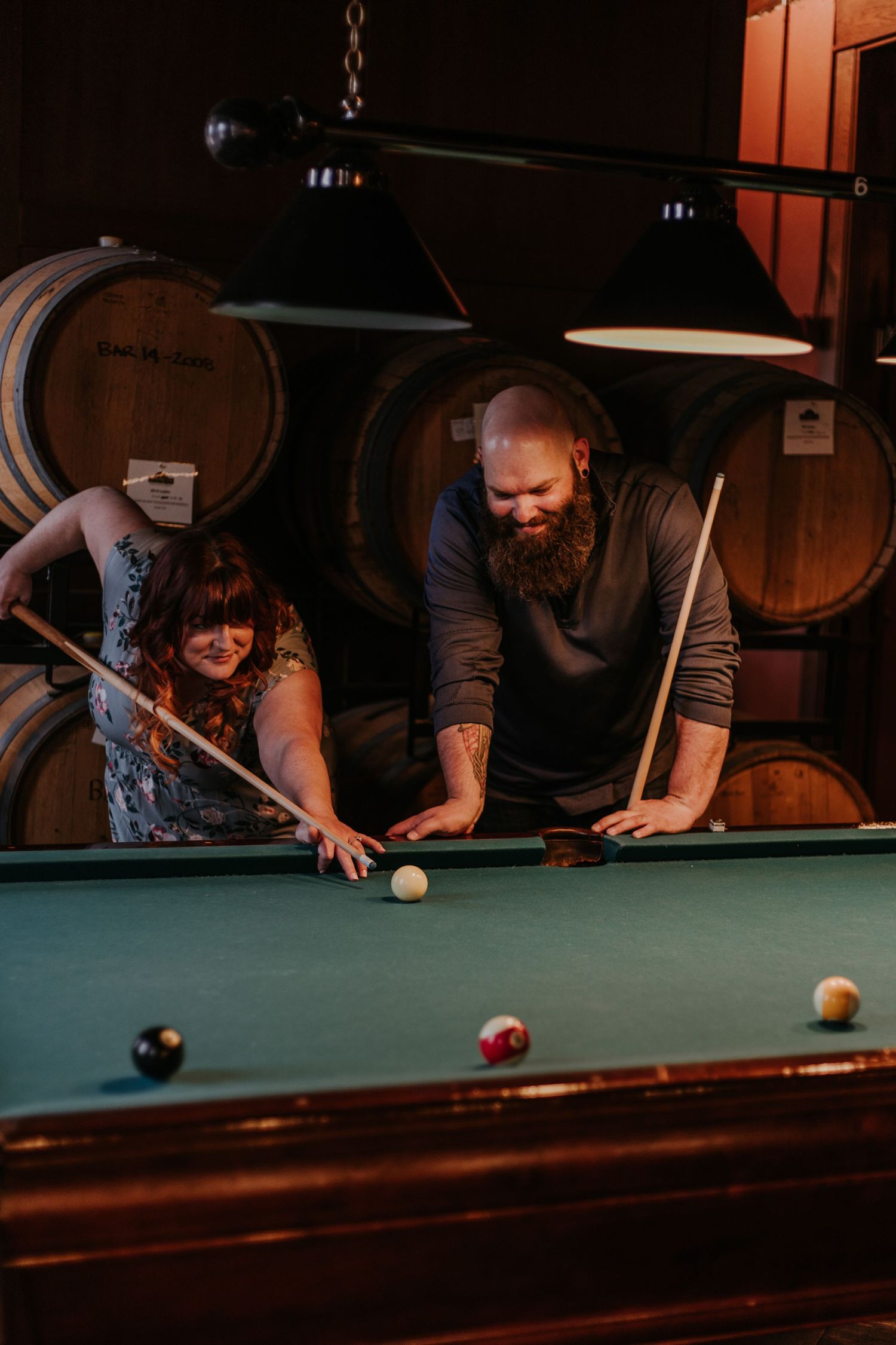 People playing pool at a dimly lit bar with wooden barrels visible in the background.