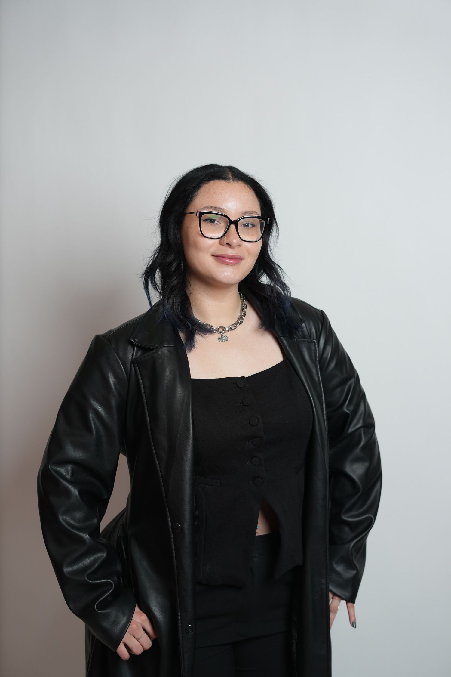 Young woman with black hair and glasses wearing all-black outfit with leather jacket, posing against light gray background.