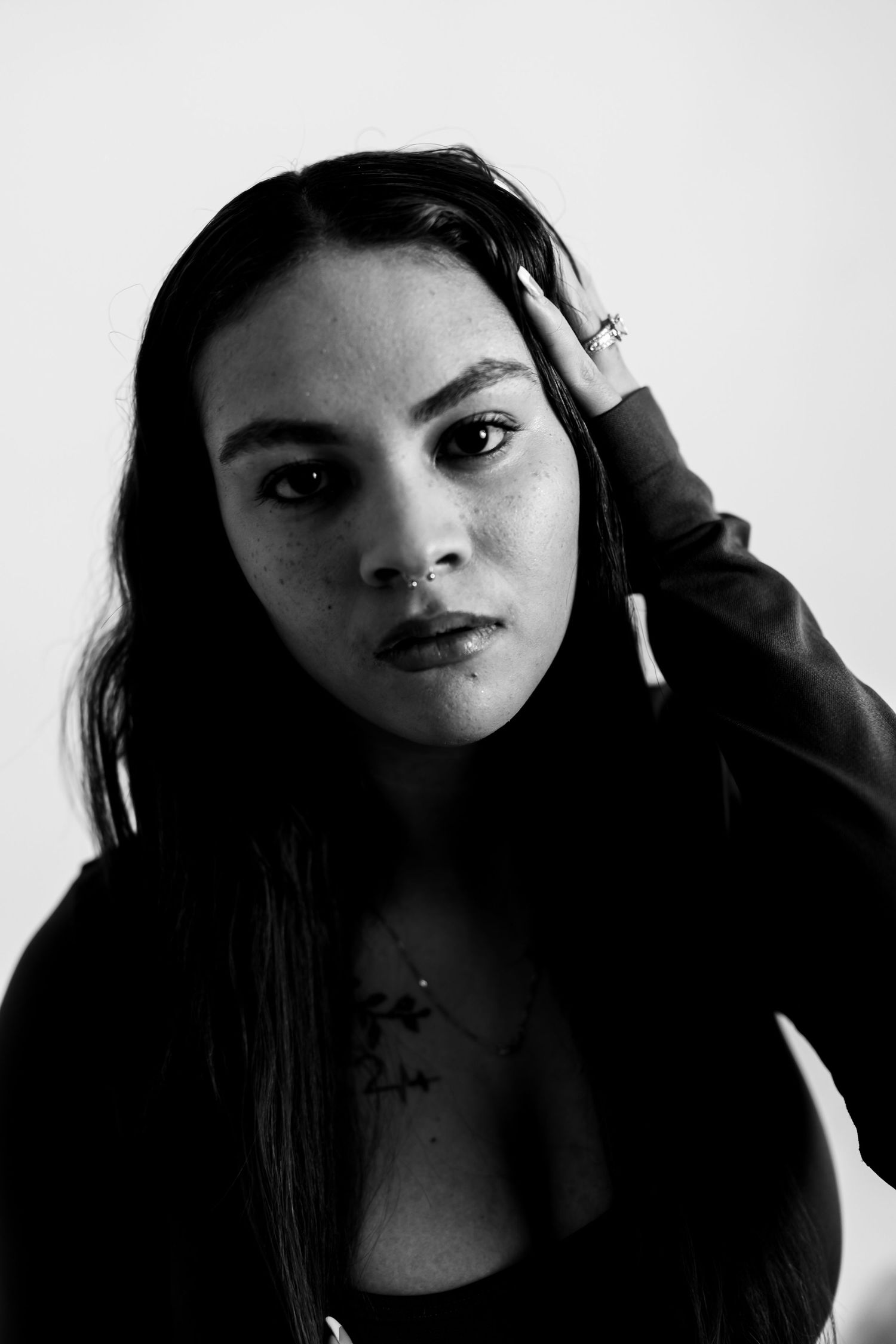 Black and white portrait of a young woman with long dark hair, freckles, and a necklace, posing with hand in hair.
