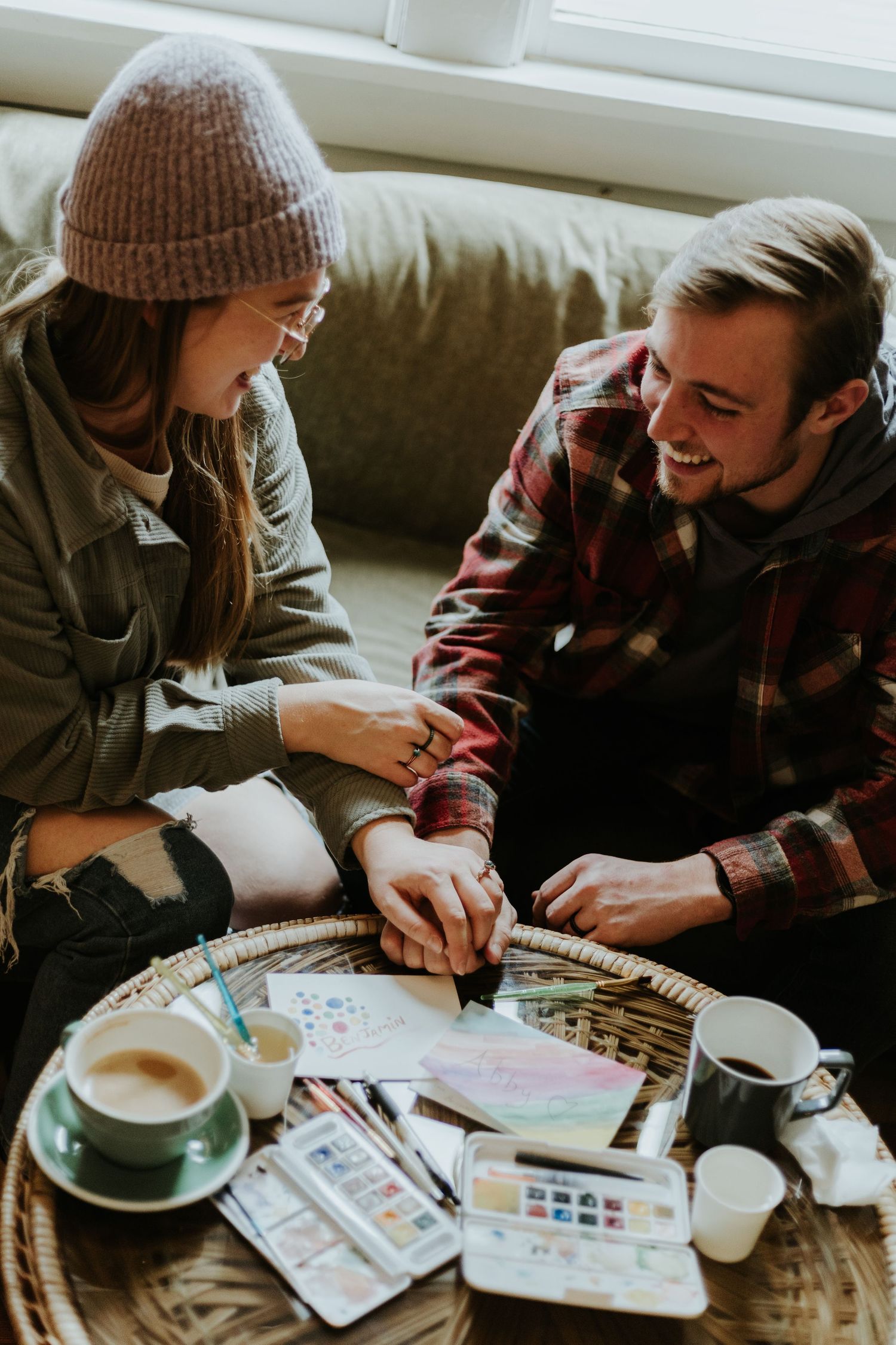 Two people sit together at a table with watercolor paints and art supplies while enjoying coffee.
