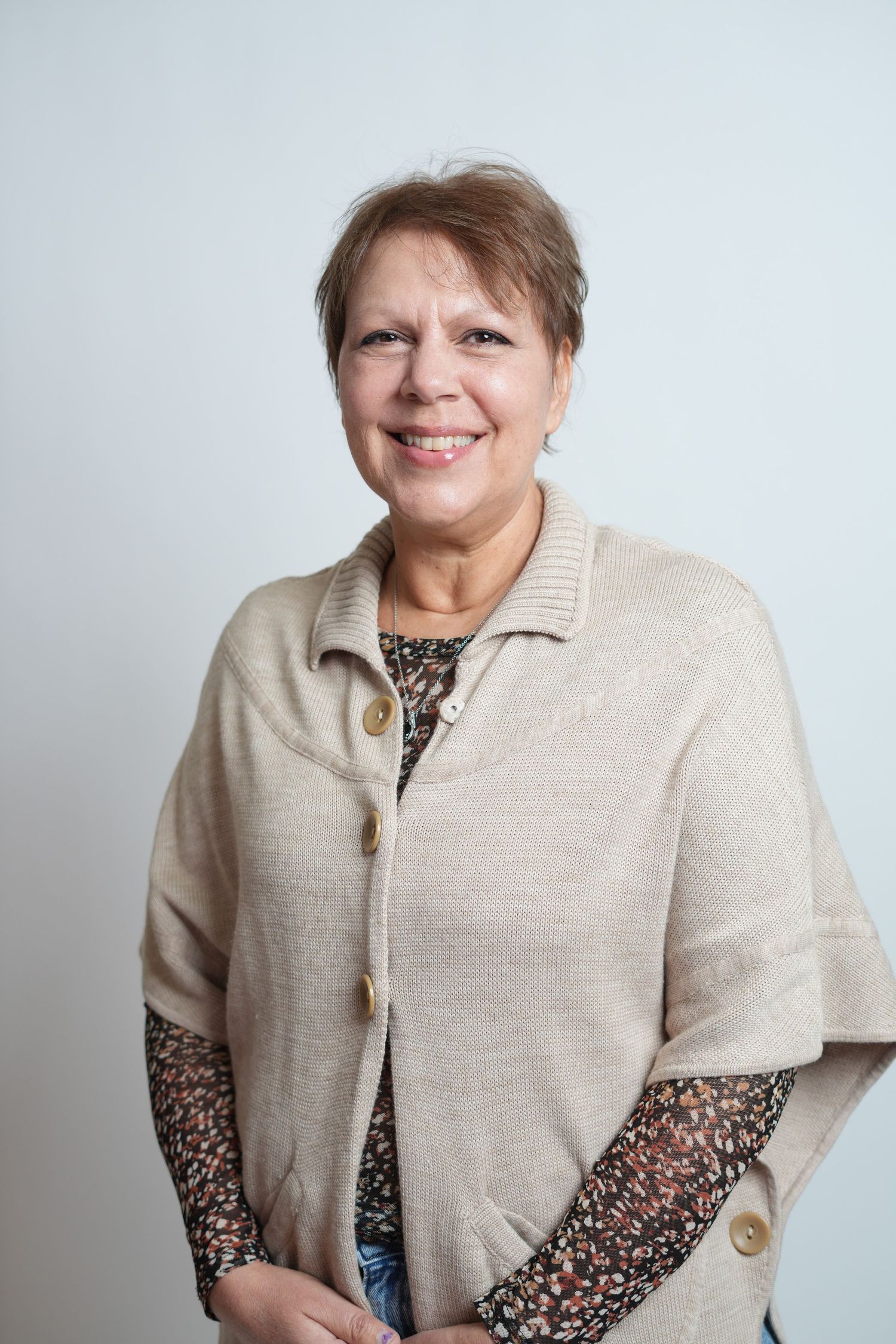 Smiling middle-aged woman with short brown hair wearing a beige knit cardigan over a floral blouse, posing against a light background.