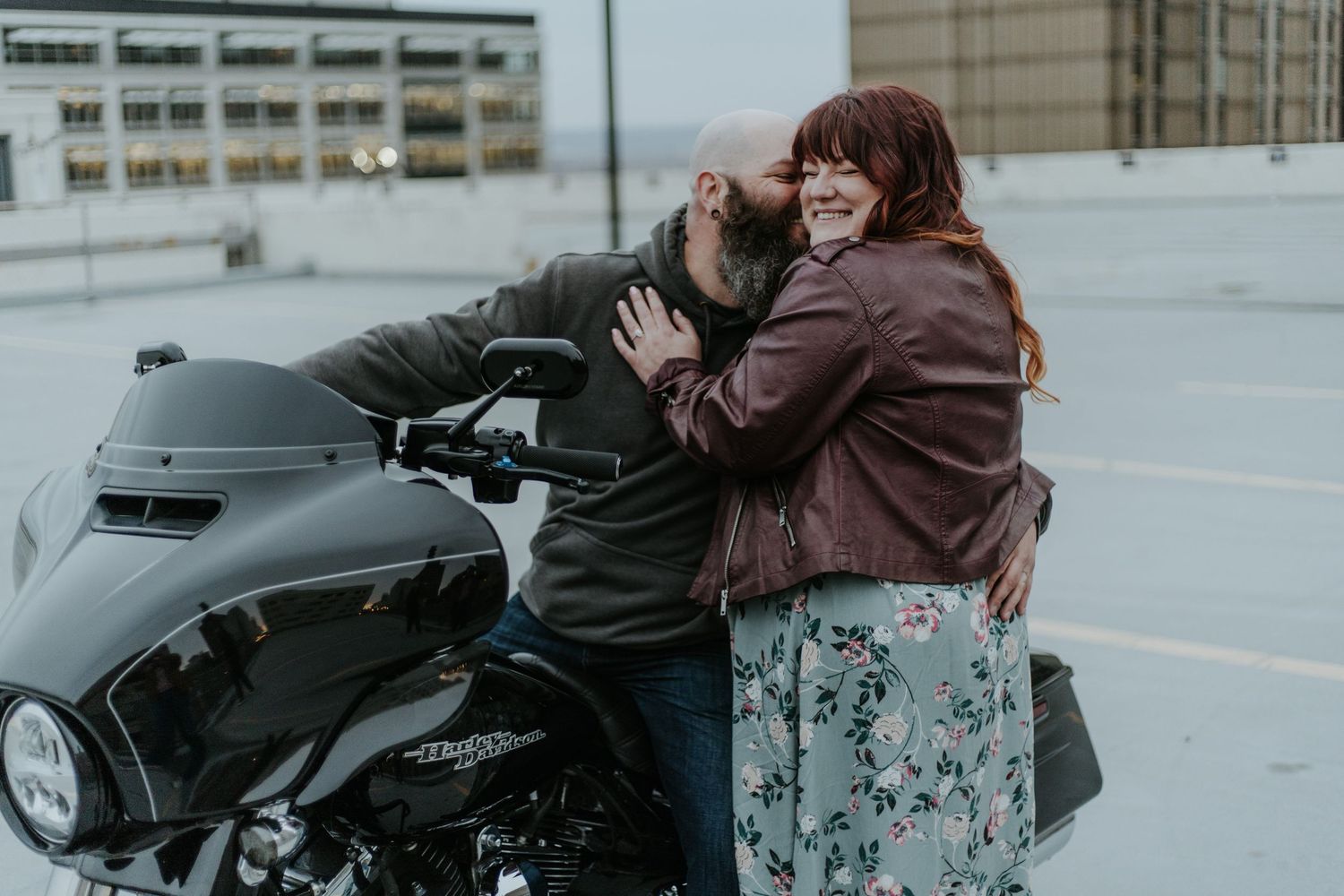 Two people share a tender moment next to a black motorcycle on a rooftop parking area.
