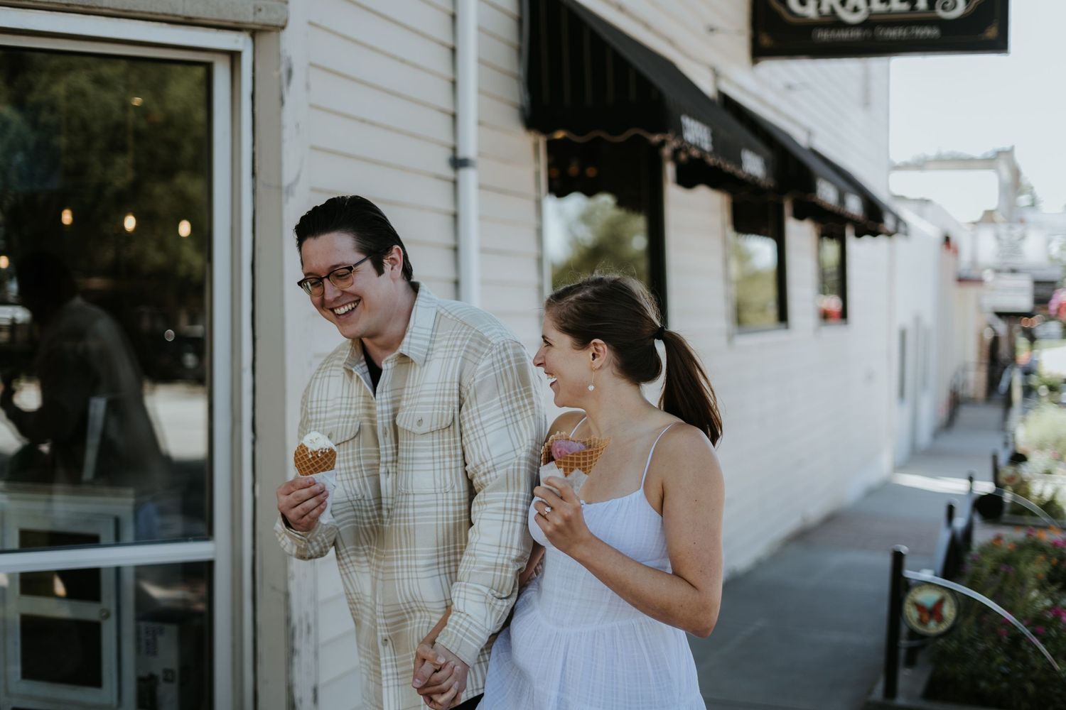A couple laughs together while enjoying ice cream cones outside an ice cream shop on a summer evening.