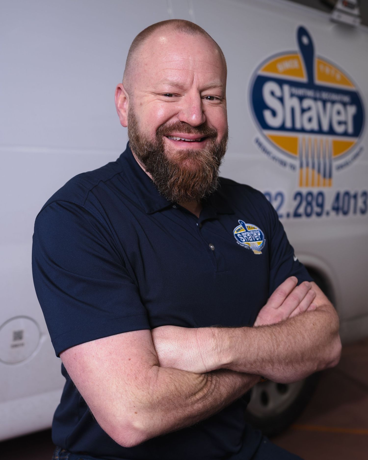 Smiling bearded man in navy Shaver Heating polo with arms crossed, posing in front of a company service van.