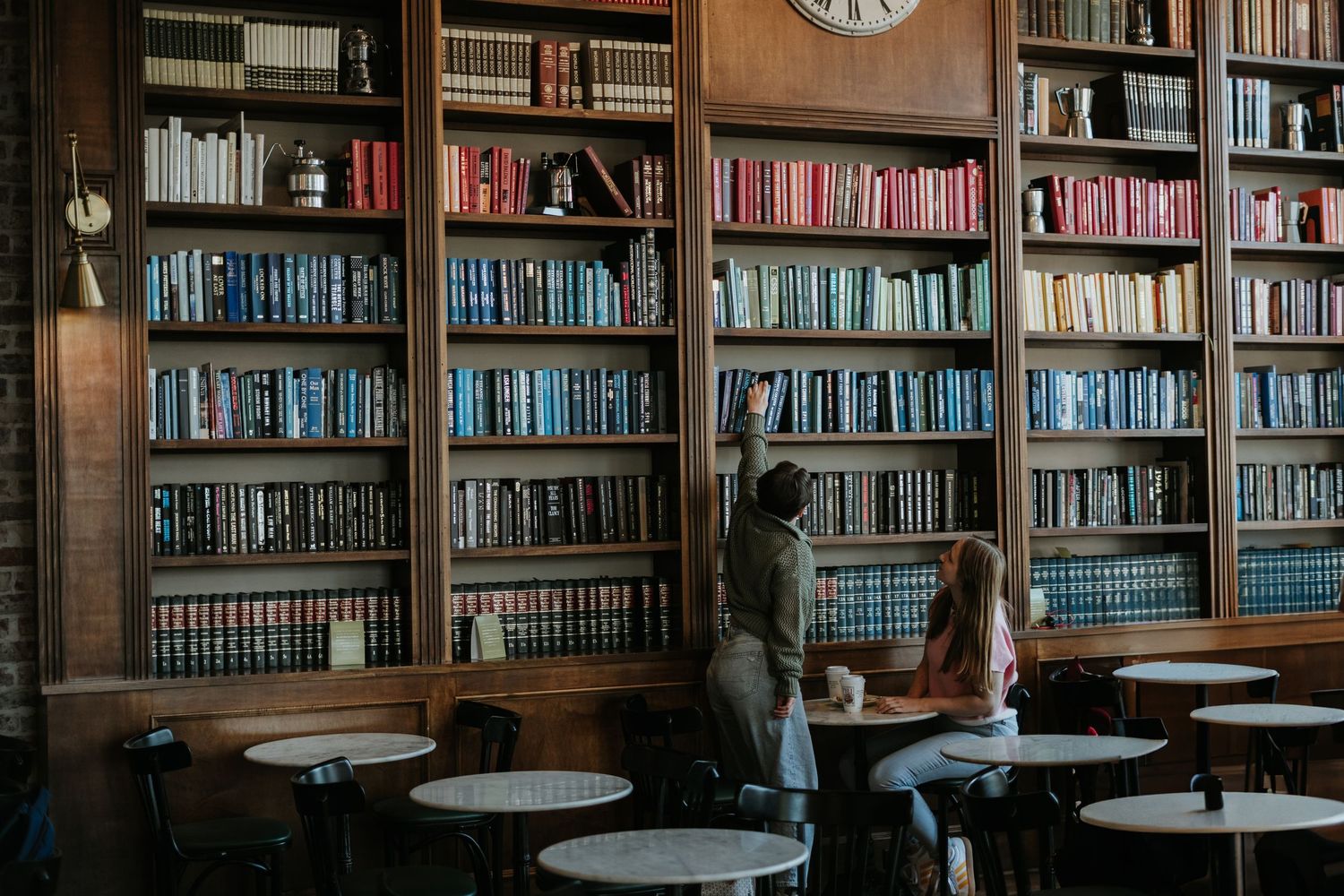 Wooden tables and chairs sit in front of towering bookshelves filled with books in a traditional library setting.