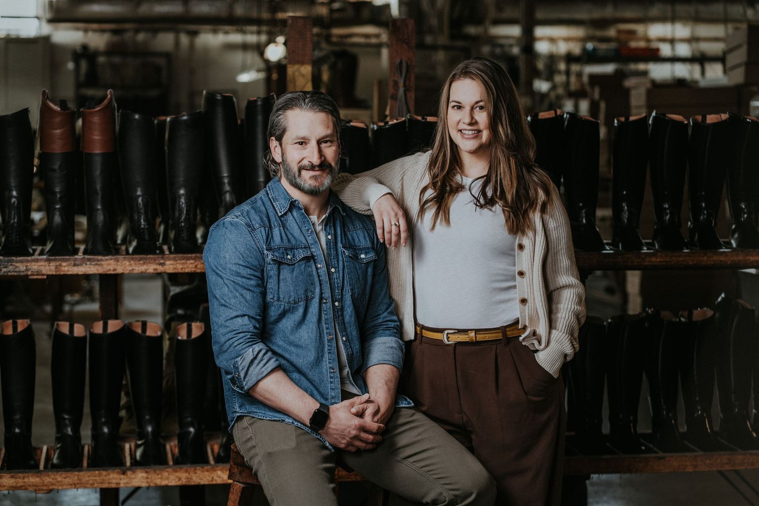 A man and woman pose smiling in a boot factory surrounded by rows of tall leather boots on display shelves.