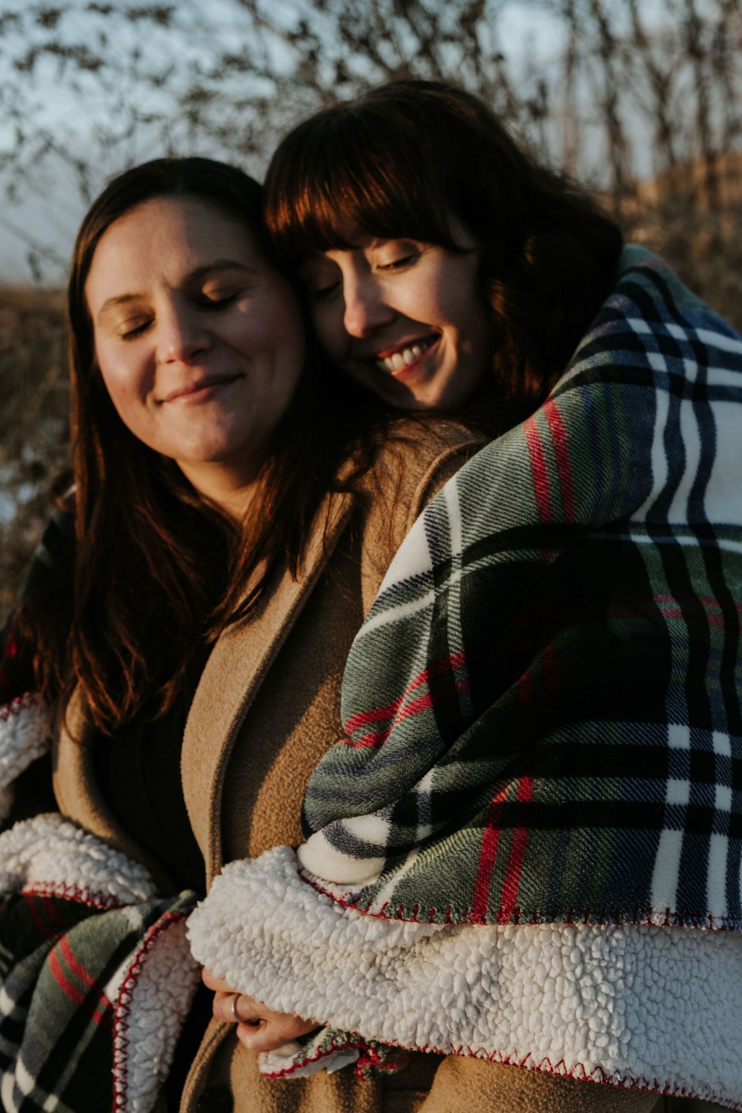 Two people share an intimate moment wrapped in a cozy plaid blanket during golden hour.