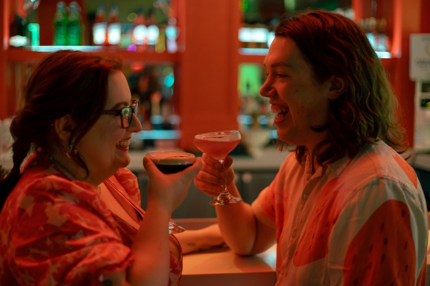 A group of people sit at a dimly lit bar counter with red ambient lighting creating a moody atmosphere.