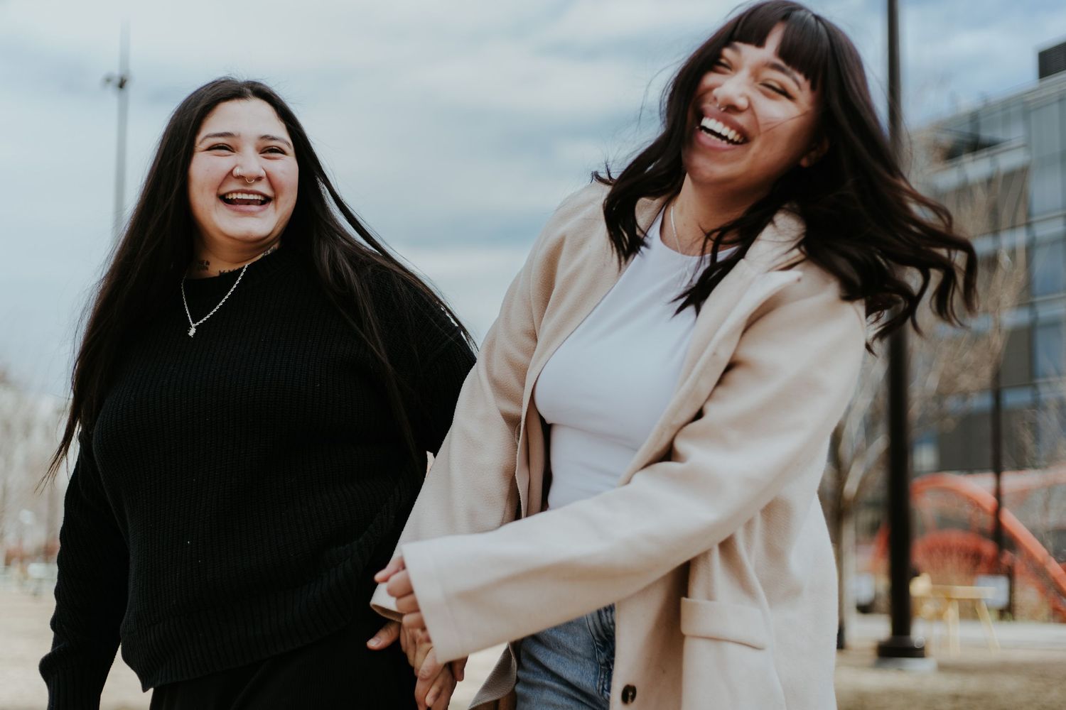 Two friends sharing joyful laughter together outdoors on a sunny day with urban architecture in the background.