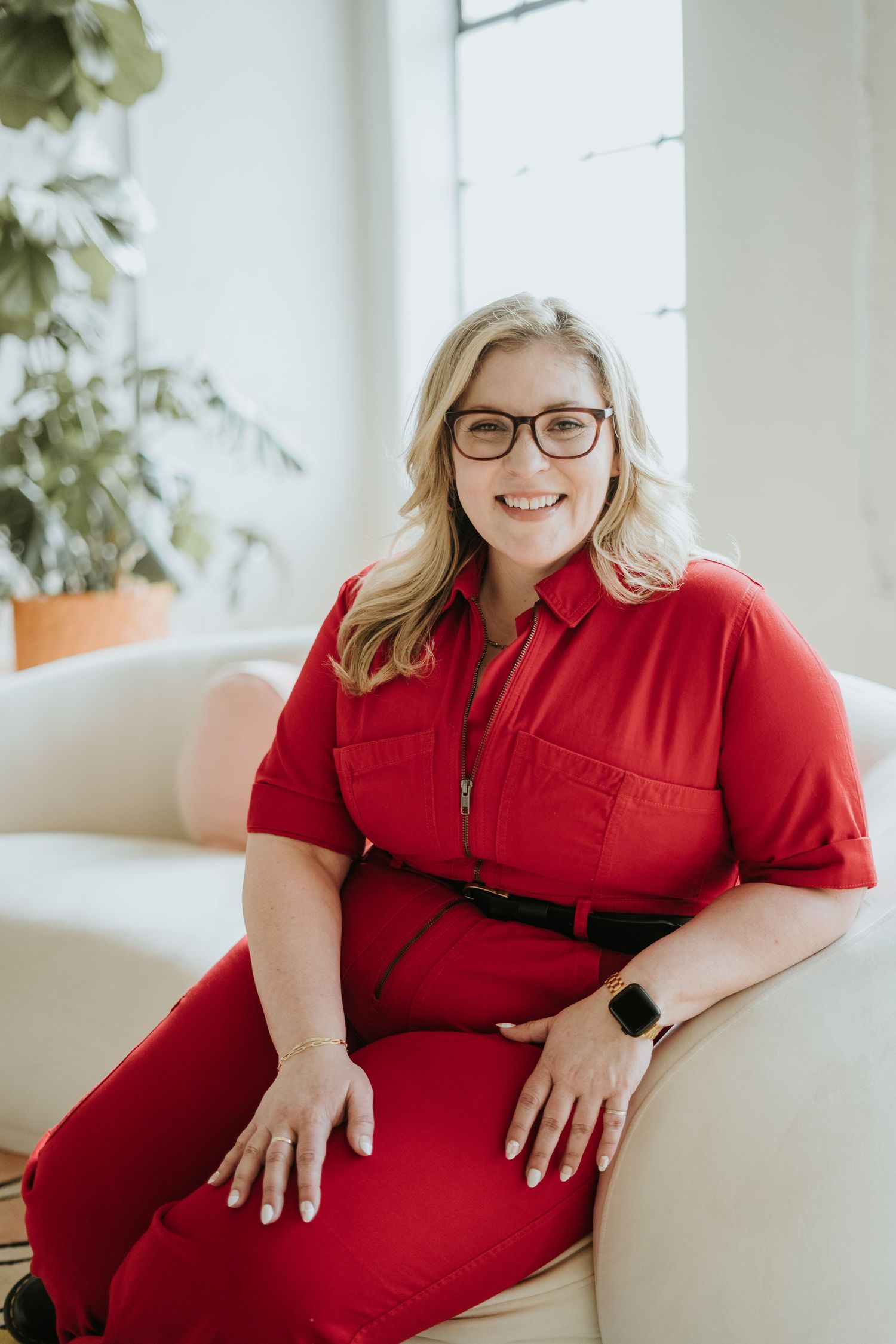 Smiling blonde woman in red jumpsuit with glasses sits on white sofa in bright modern space with plants.