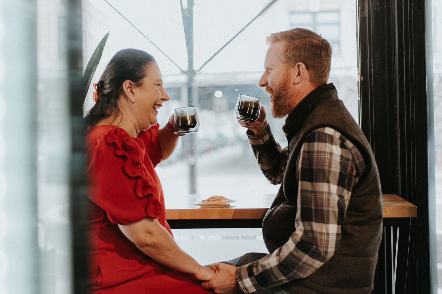 A couple shares a laugh while drinking wine together by a window on a winter day.