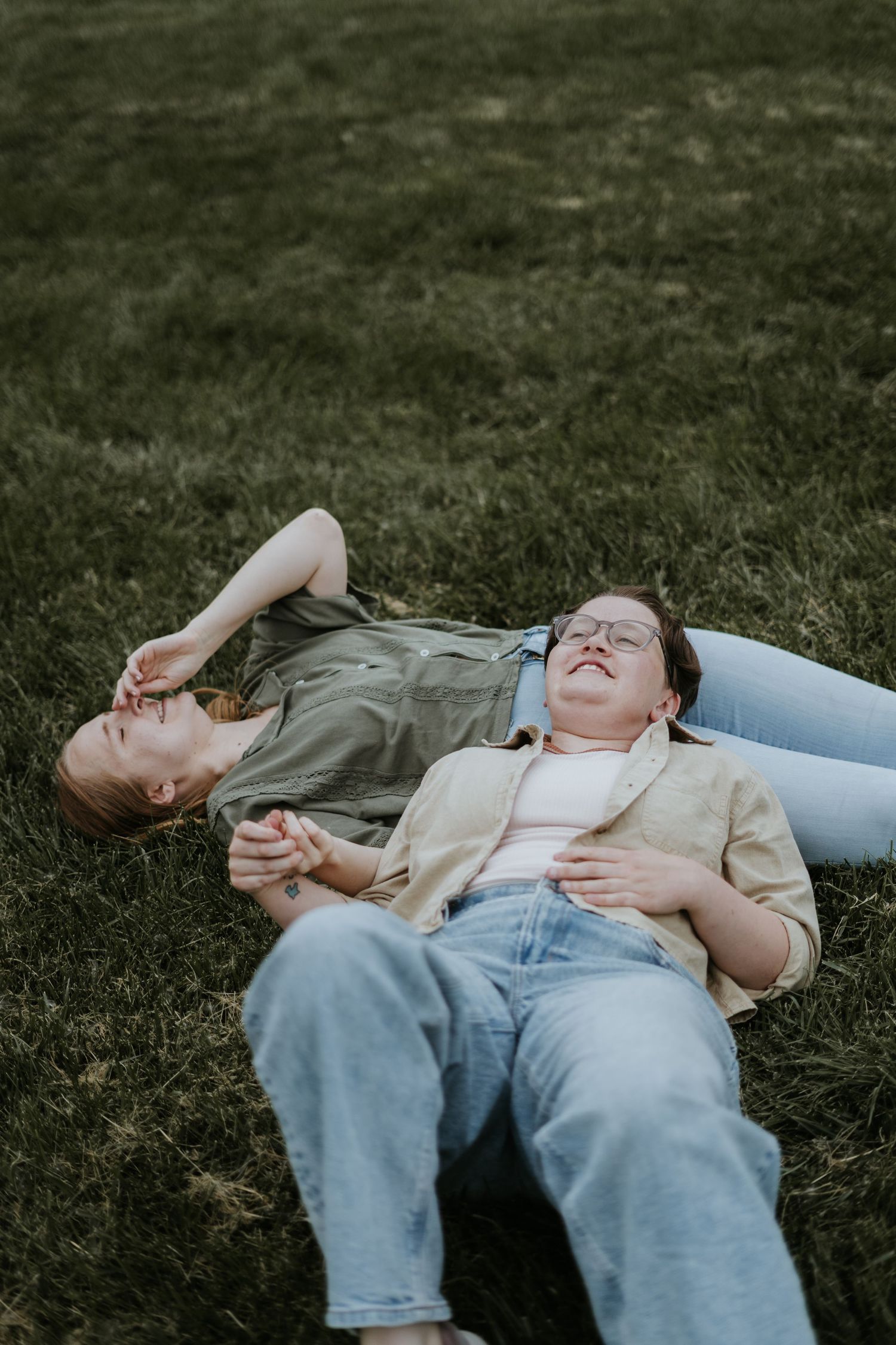 Two people laying in the grass together on a sunny day wearing casual denim and earth-toned clothing.