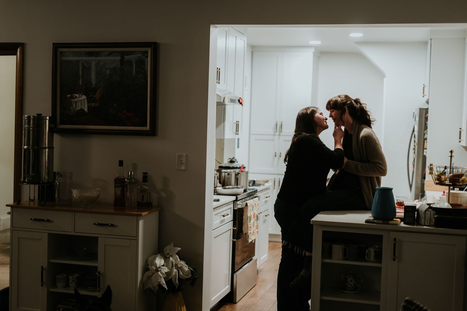 A couple shares an intimate moment in a dimly lit kitchen with white cabinets and wooden floors.
