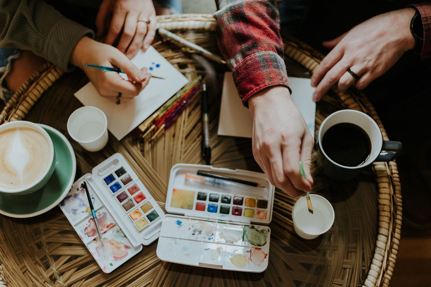 Overhead view of hands painting with watercolors while enjoying coffee and lattes on a wooden table.