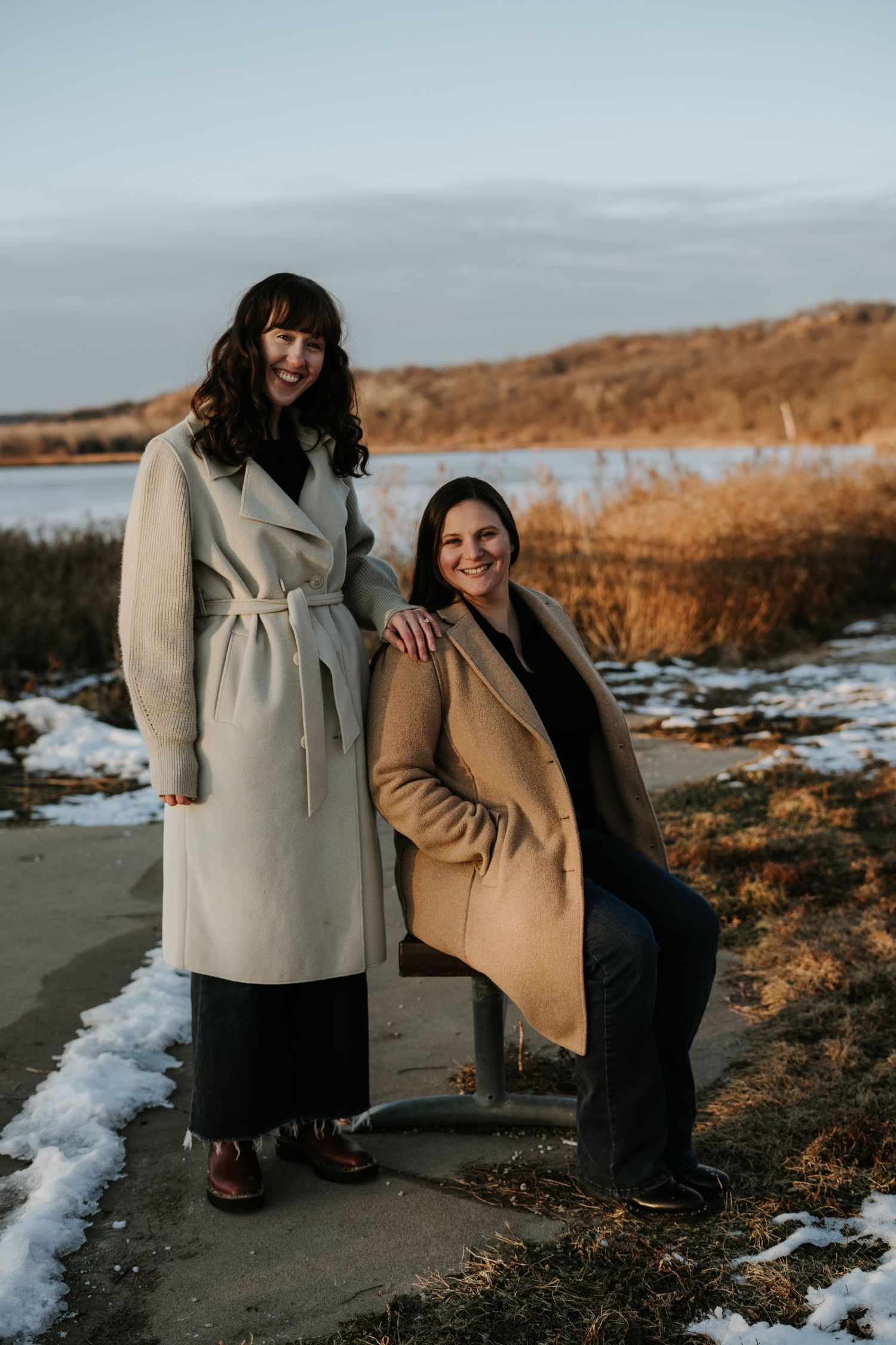A couple poses together outdoors in winter, wearing warm coats against a backdrop of snow and golden light.