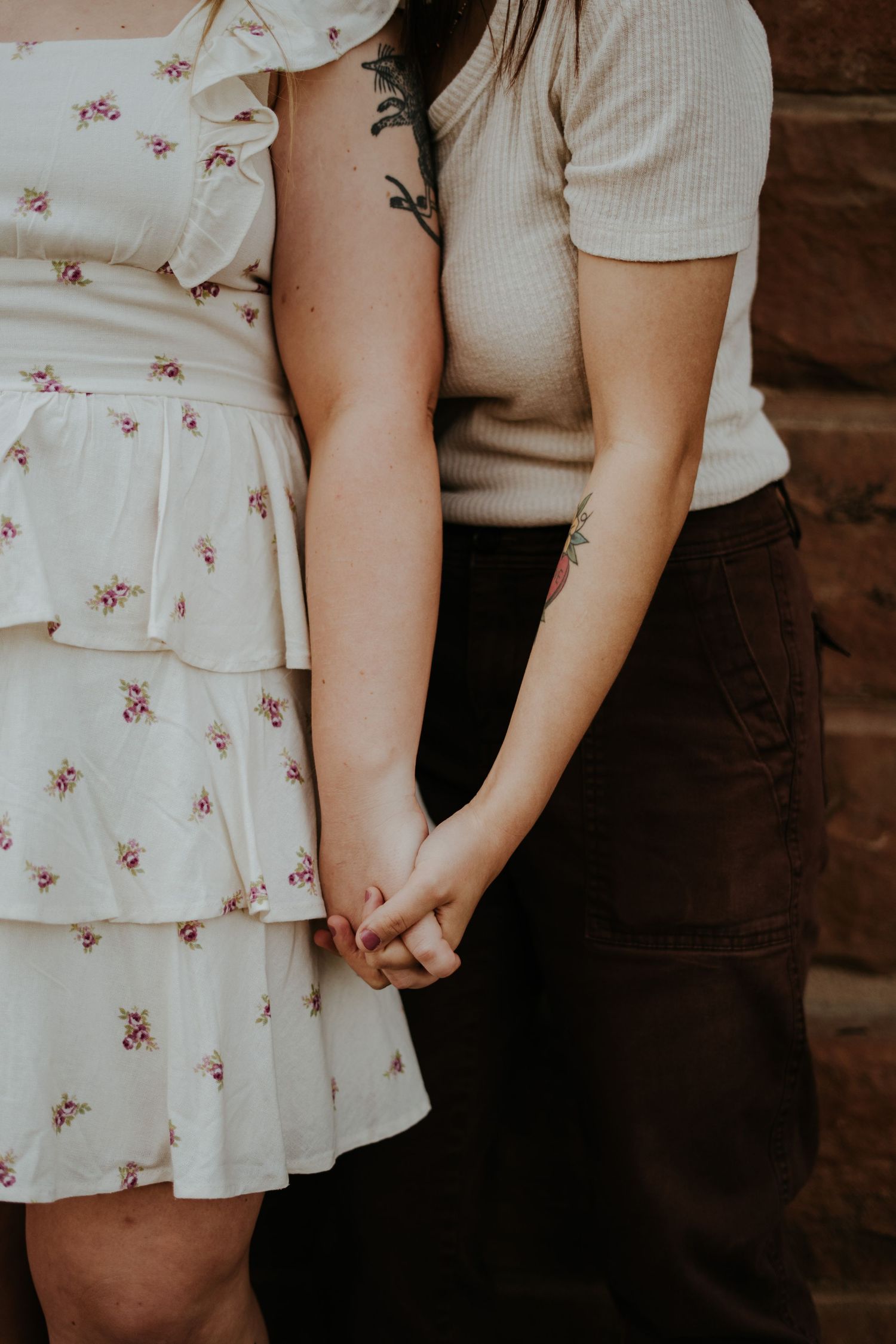 Close-up of couple holding hands, wearing floral dress and casual attire against brick wall in romantic pose.