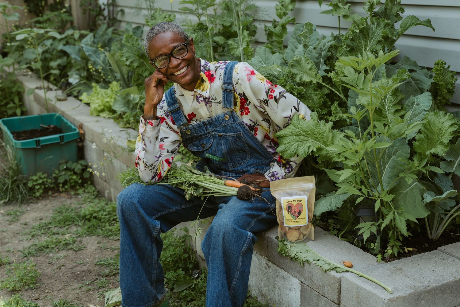 Smiling person in overalls sits in a lush garden holding fresh vegetables and a seed packet.
