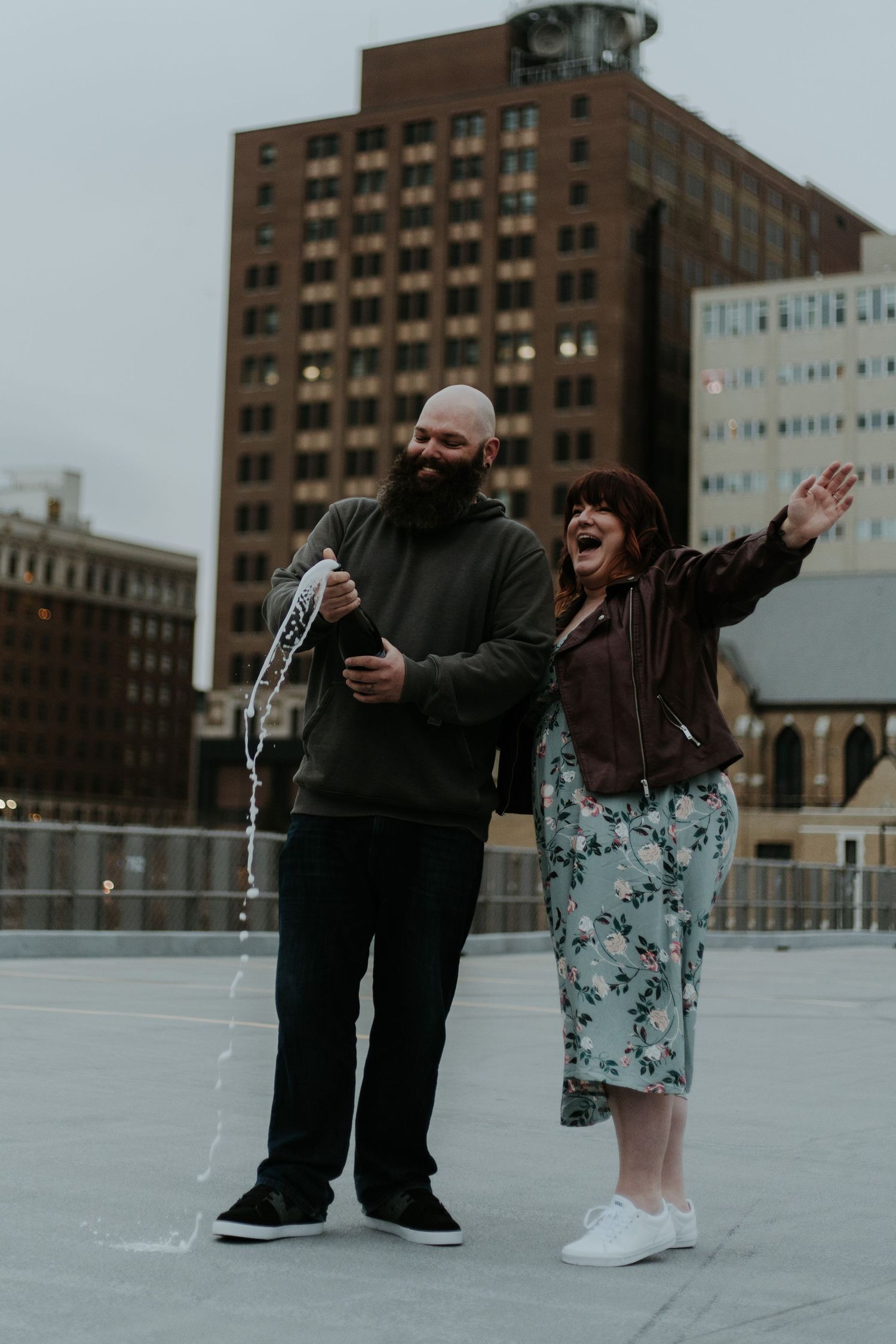 A couple laughs and dances together on an urban rooftop with tall buildings in the background on an overcast day.