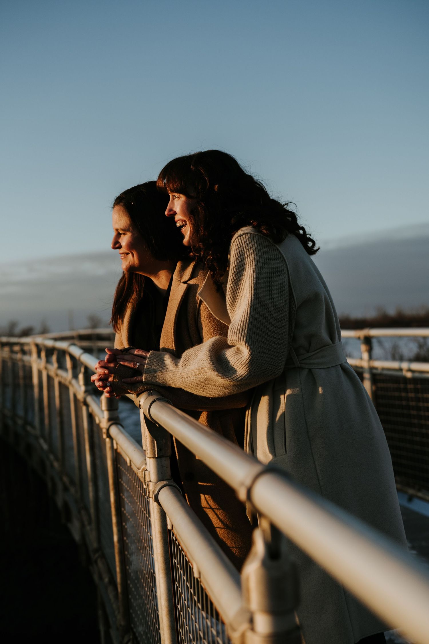 Two people lean against a wooden railing at sunset, creating silhouettes against the dusky sky.