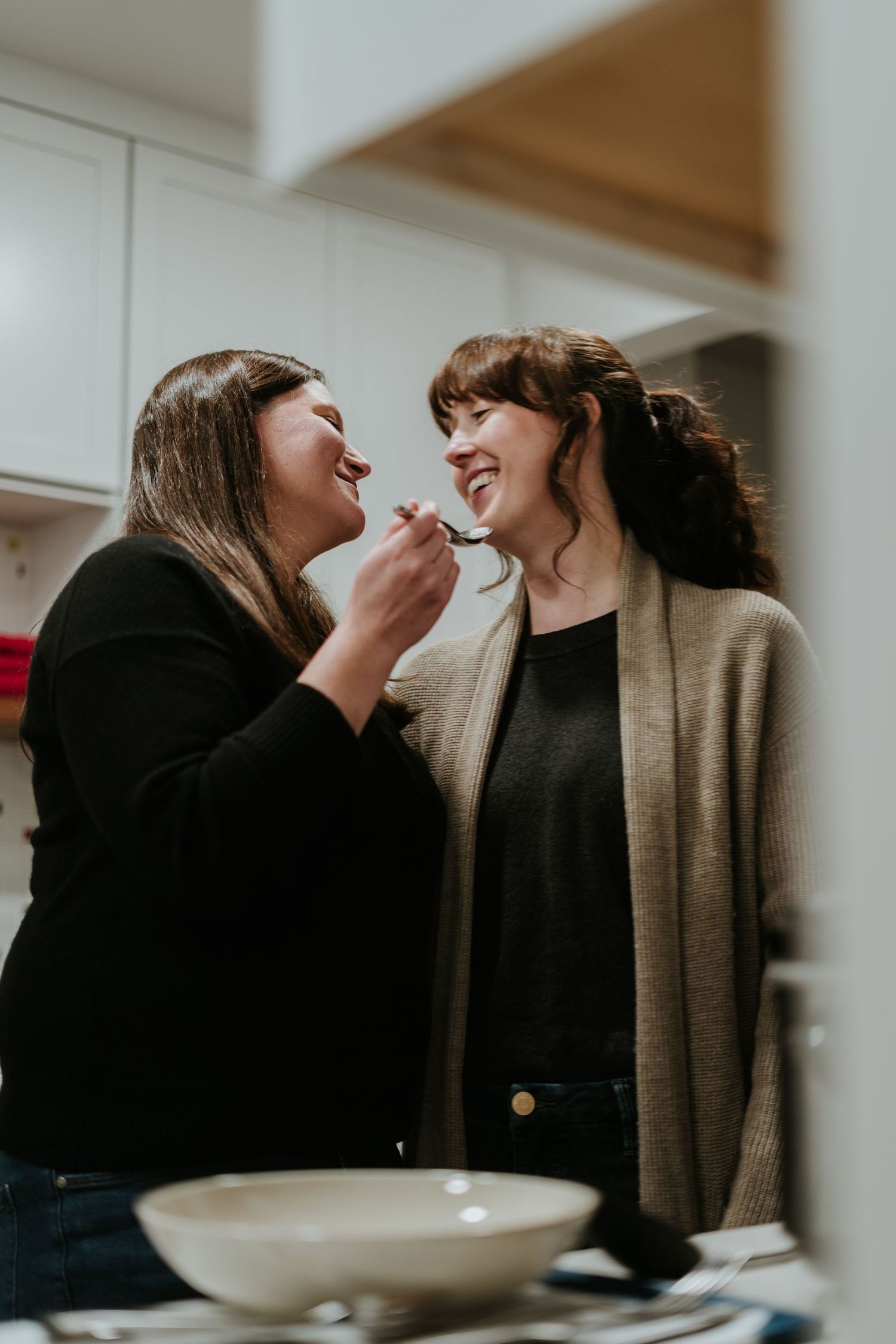Two people in dark clothing and beige sweaters share an intimate moment while eating from a white bowl.