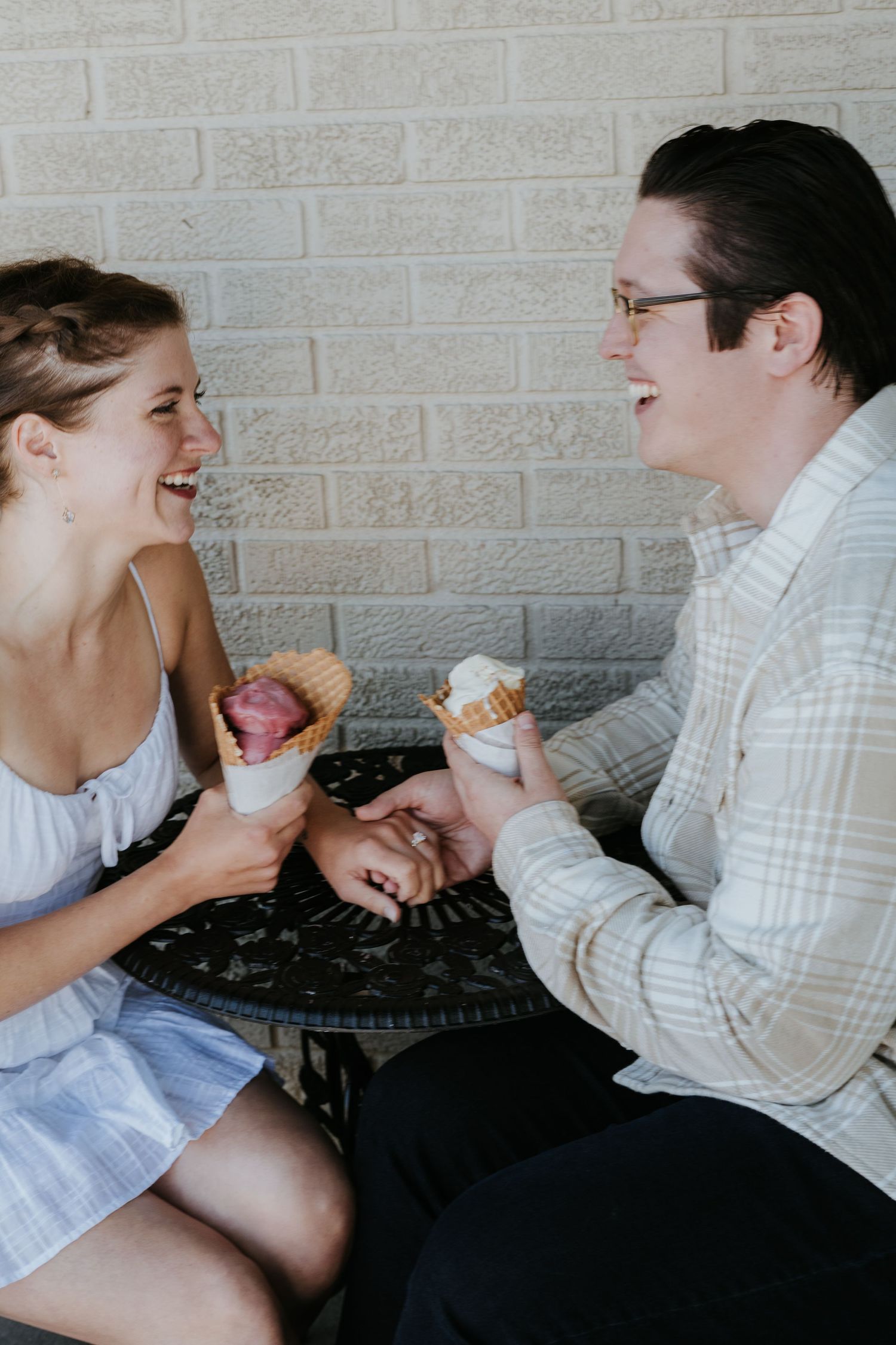 A couple sitting together outdoors share ice cream cones while laughing and enjoying each other's company.