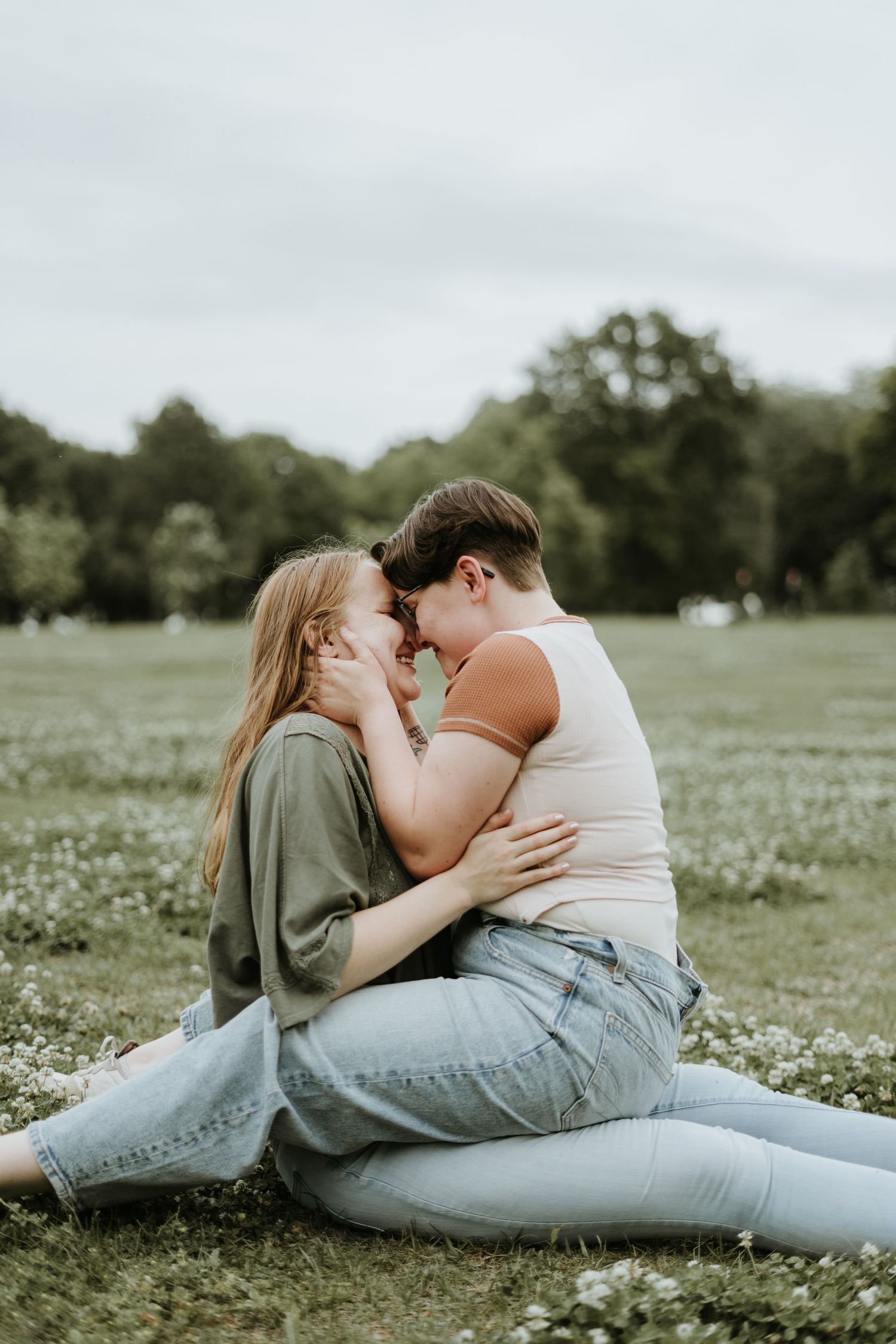 A couple lies together in a grassy field filled with small white flowers on a spring day.