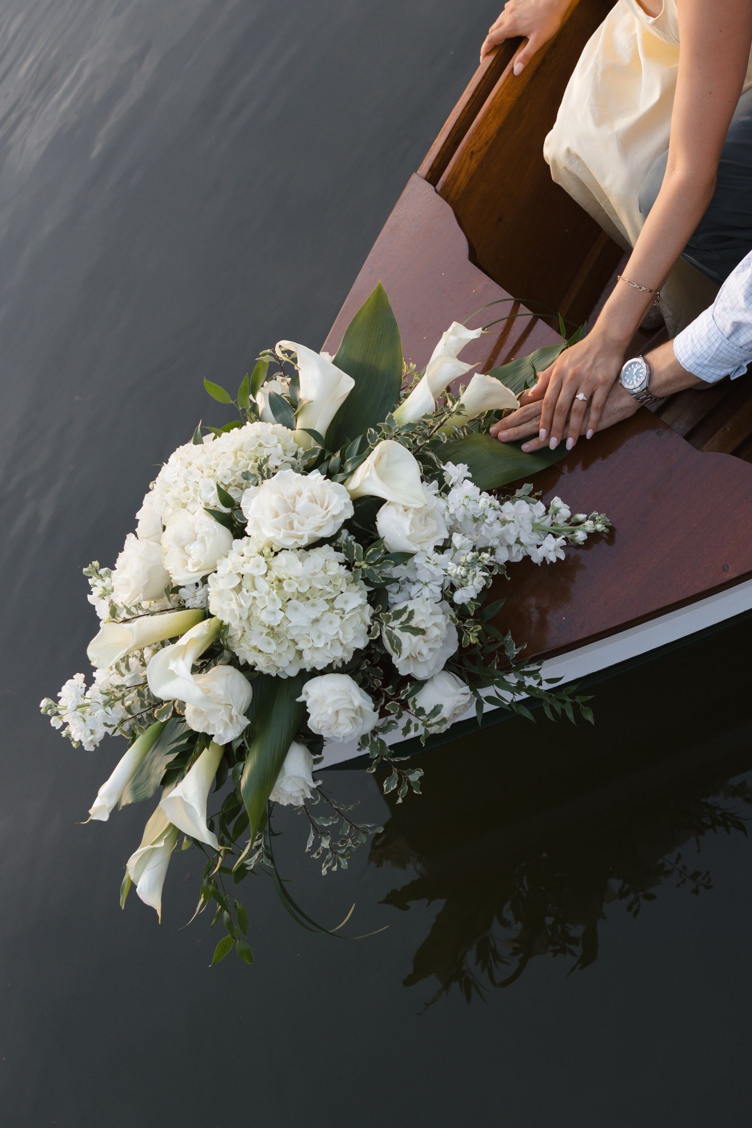 Elegant white wedding bouquet with hydrangeas and calla lilies on a wooden boat bow, with couple's hands visible.