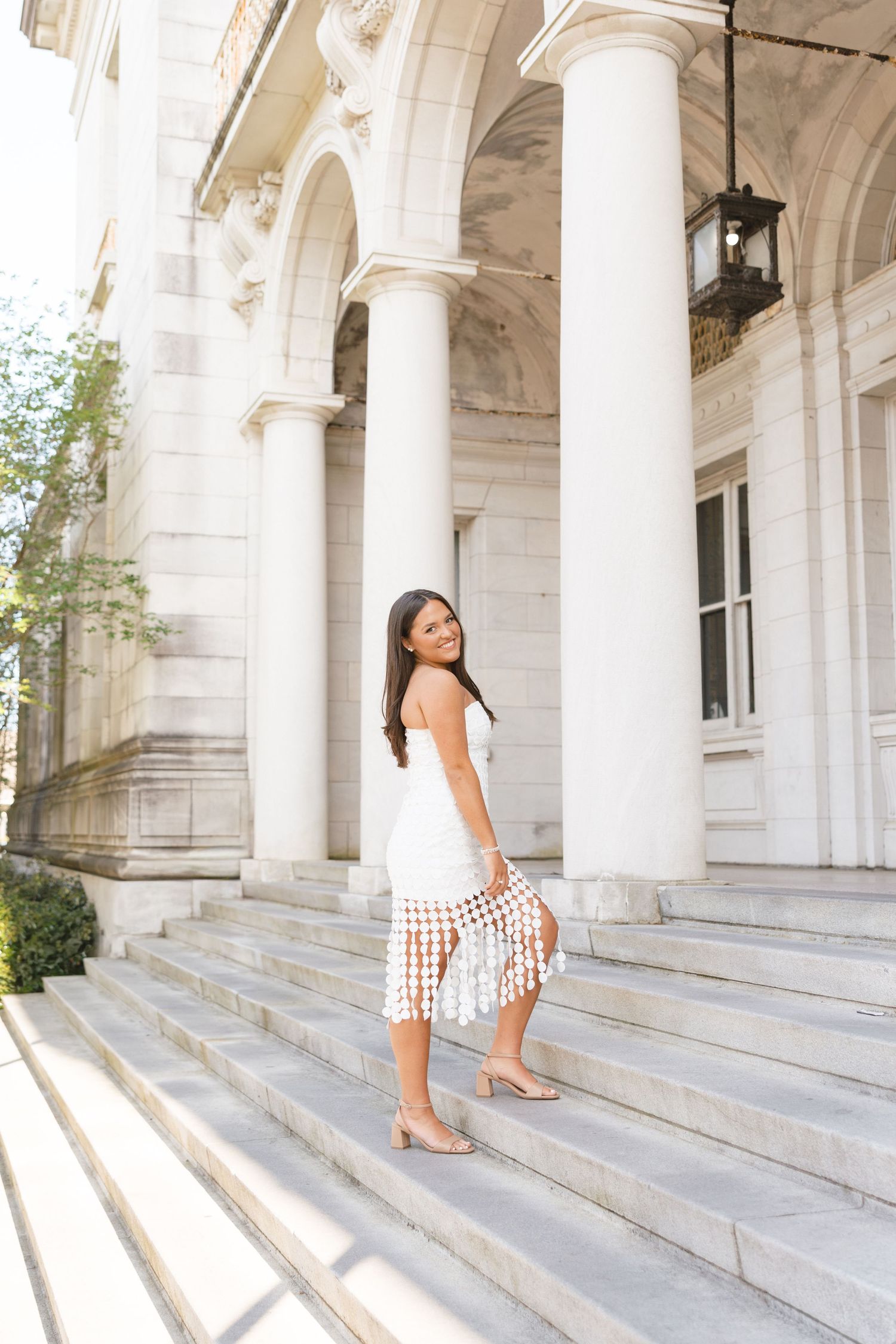 Brunette woman in white fringed midi dress posing on marble steps of grand building.