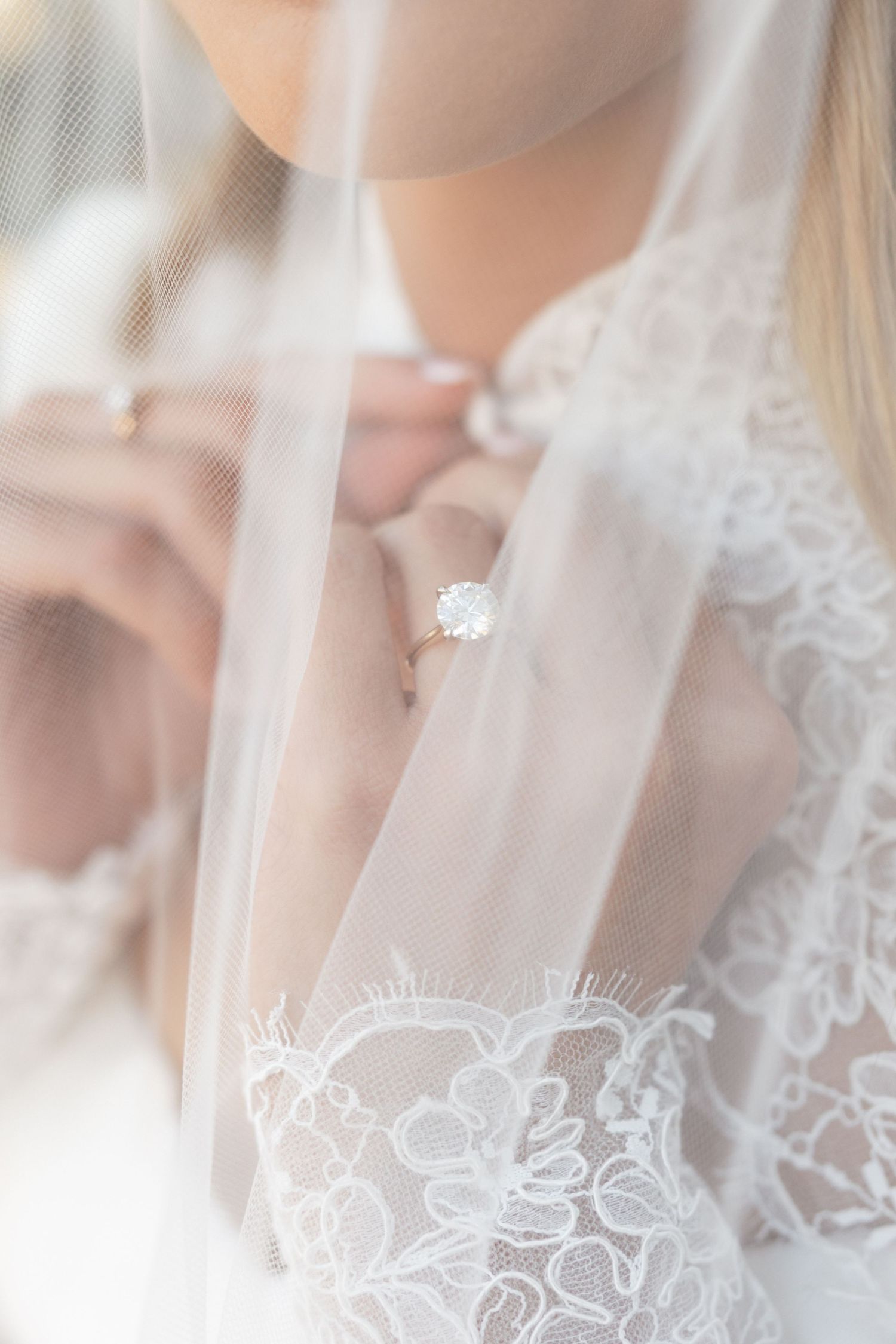 Close-up of oval solitaire engagement ring on bride wearing lace wedding gown with veil.