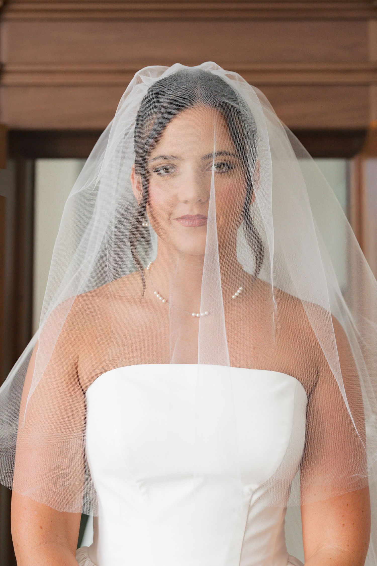 Beautiful dark-haired bride wearing strapless white gown, pearl necklace, and flowing wedding veil.