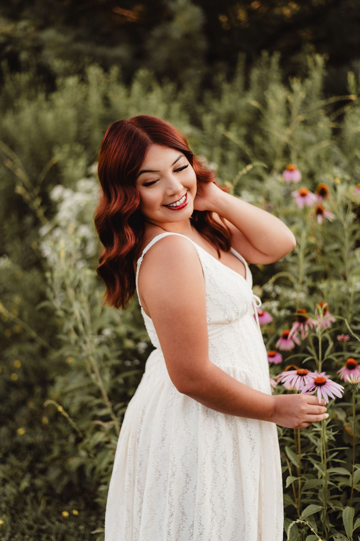 Woman in white lace dress smiling among wildflowers holding a pink echinacea bloom in a lush garden.