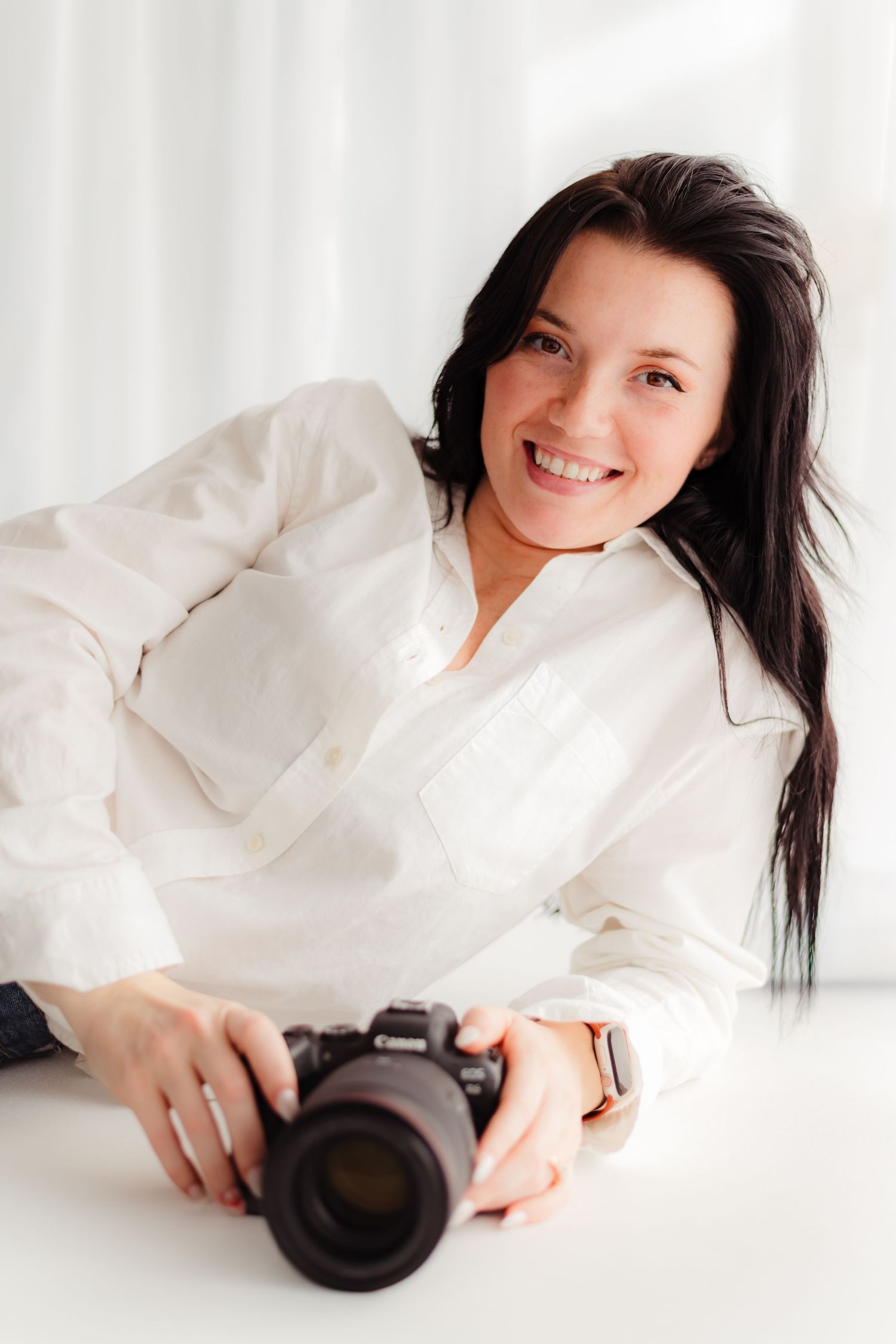 Smiling brunette woman in white shirt holding a Canon DSLR camera while leaning on a white surface.