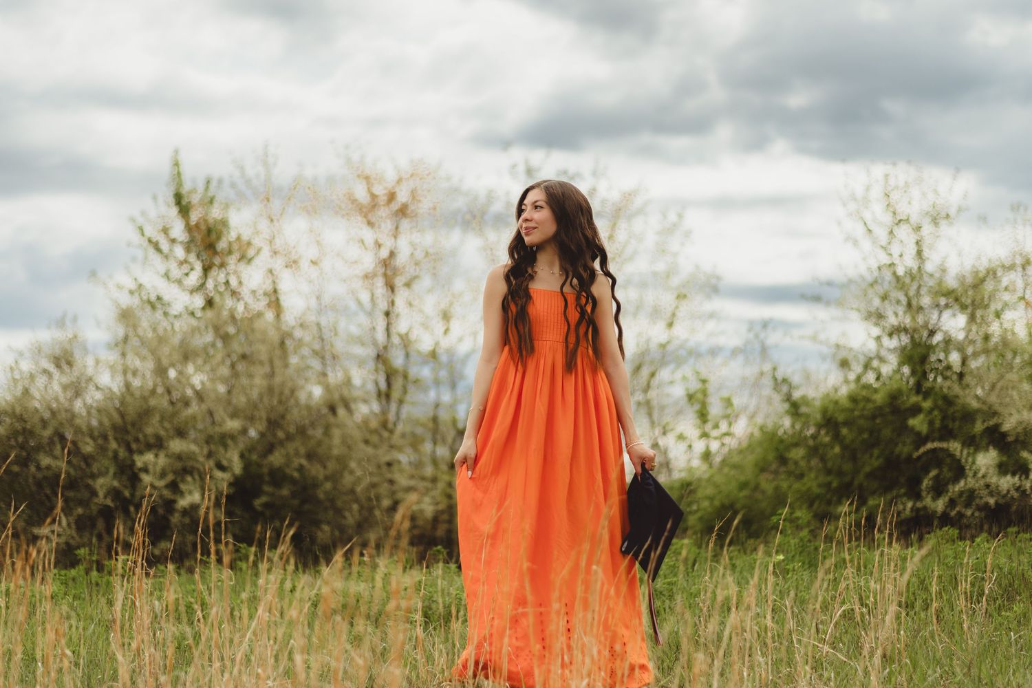 A figure in a flowing orange dress stands gracefully among tall grass in a dreamy outdoor setting.