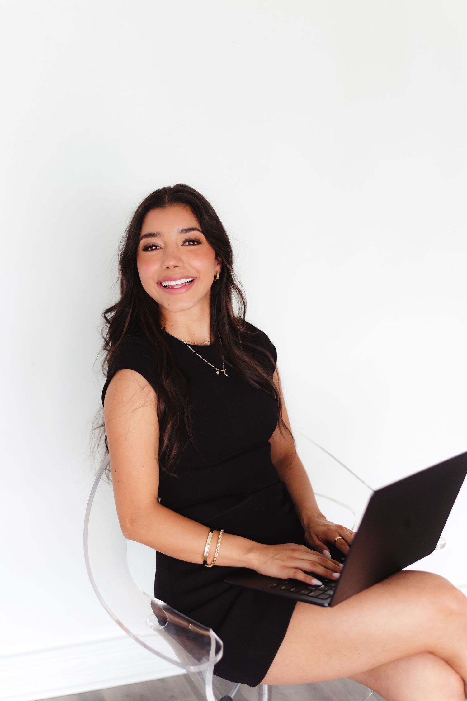 Young woman in black dress sitting on clear chair with laptop, smiling confidently against white background.