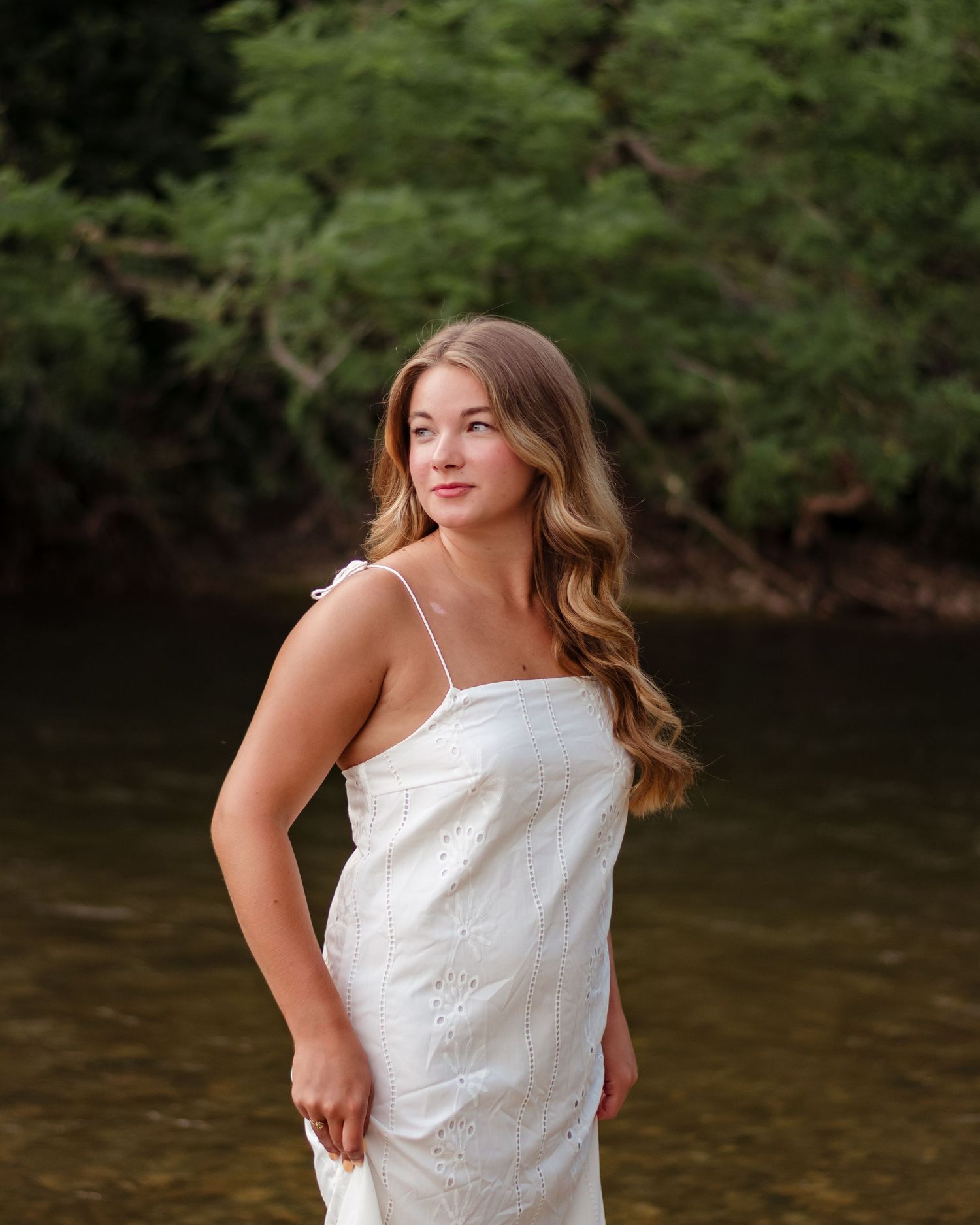 Senior in white sundress poses by a river bank during a summer portrait session in nature in Lafayette, Indiana.