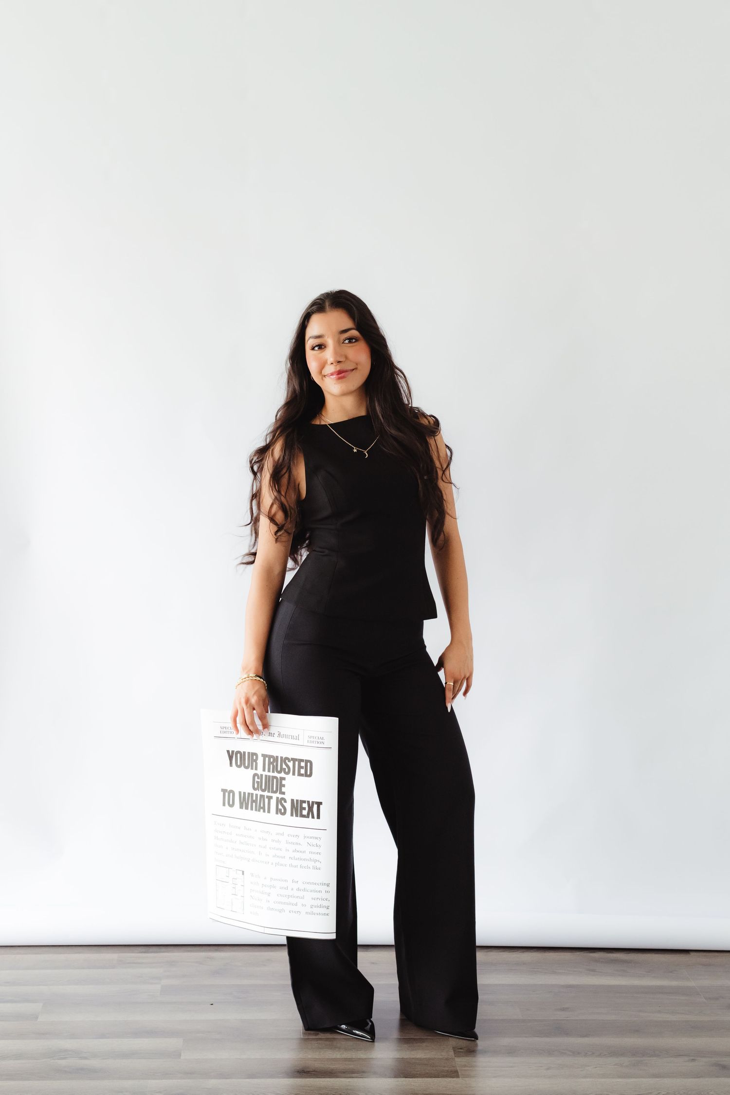 Woman in black outfit holding a 'Your Trusted Guide to What is Next' sign against a white wall.