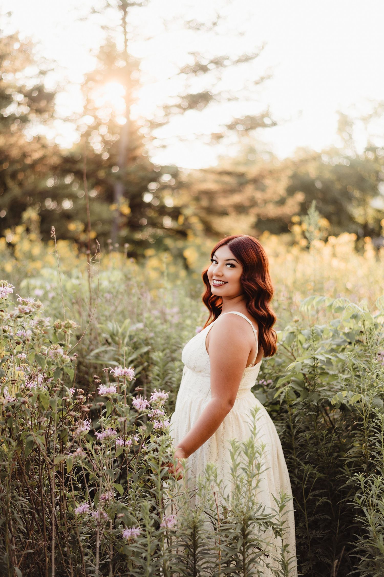 Smiling woman in white dress standing among wildflowers in a sunlit garden.