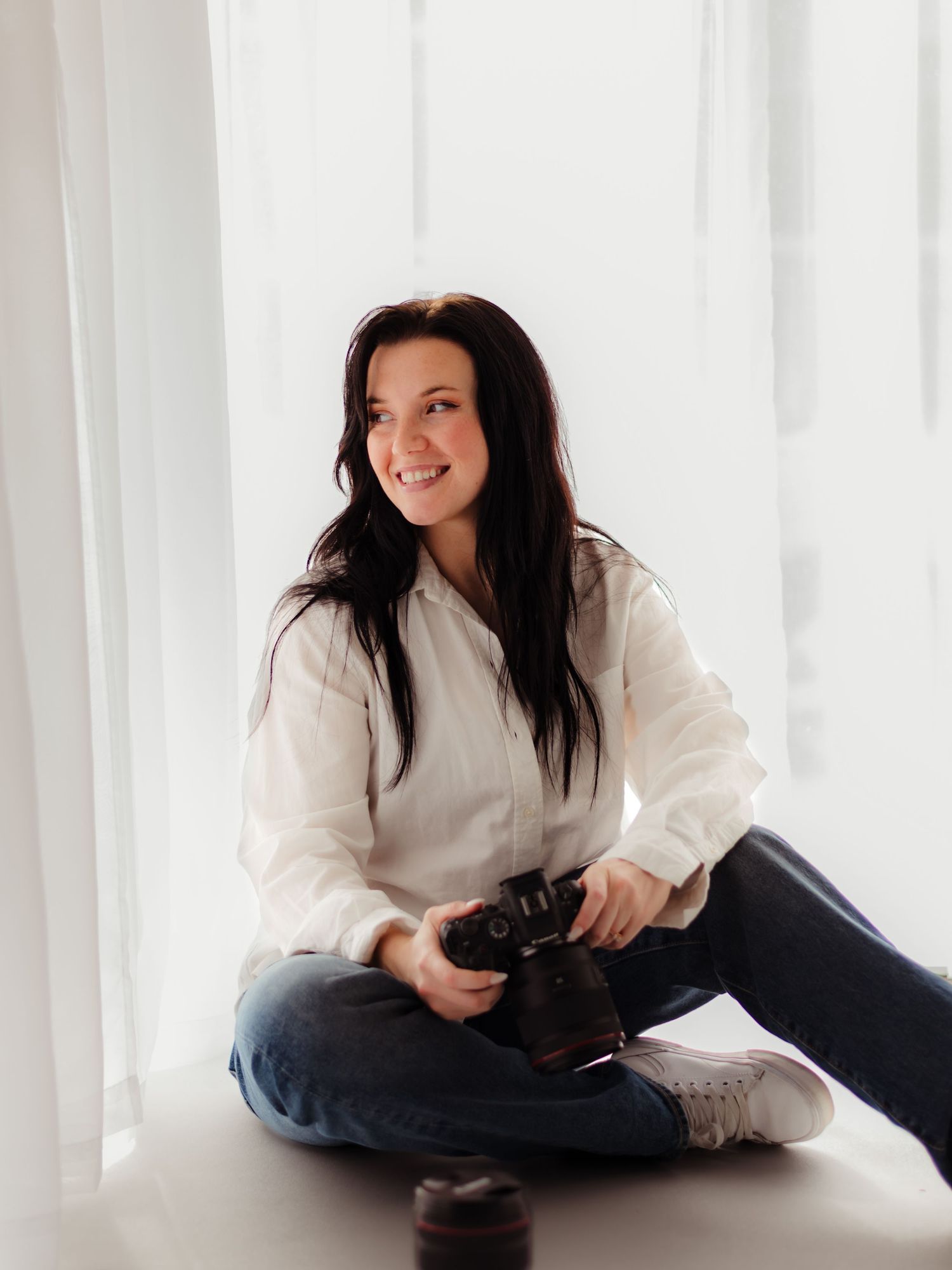 Woman with dark hair sits cross-legged holding a camera, smiling in a bright white room wearing a white shirt and jeans.
